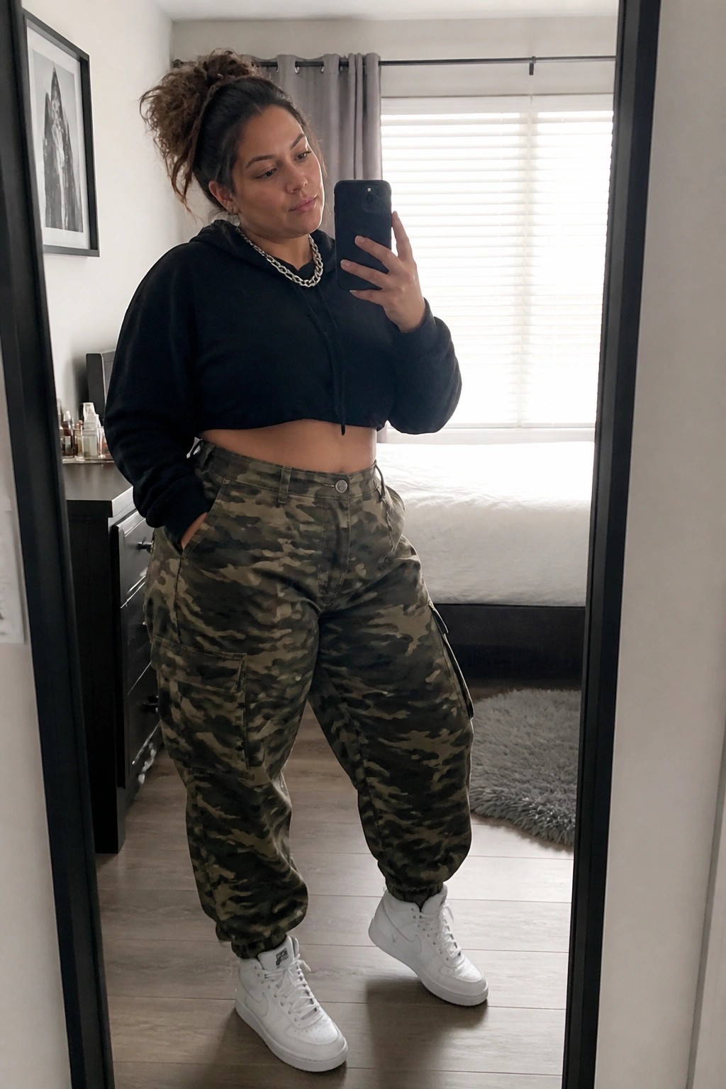 Woman in black cropped hoodie and camouflage cargo pants taking bedroom mirror selfie.