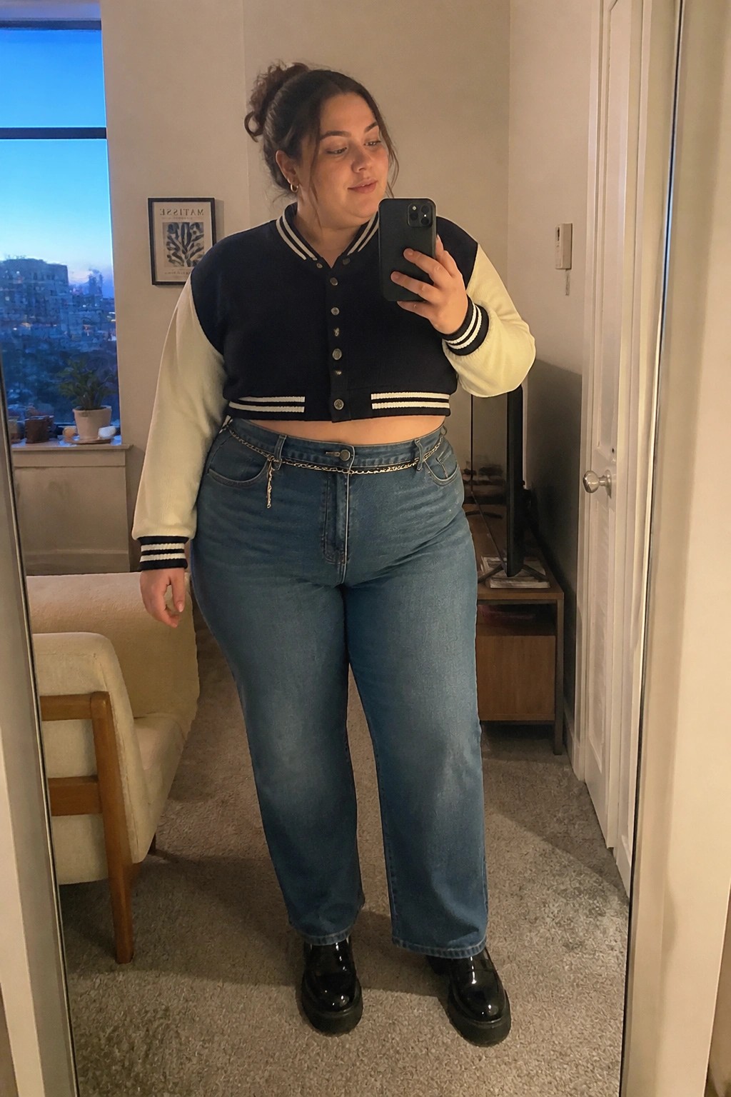 Woman in cropped navy varsity jacket and blue jeans taking mirror selfie.