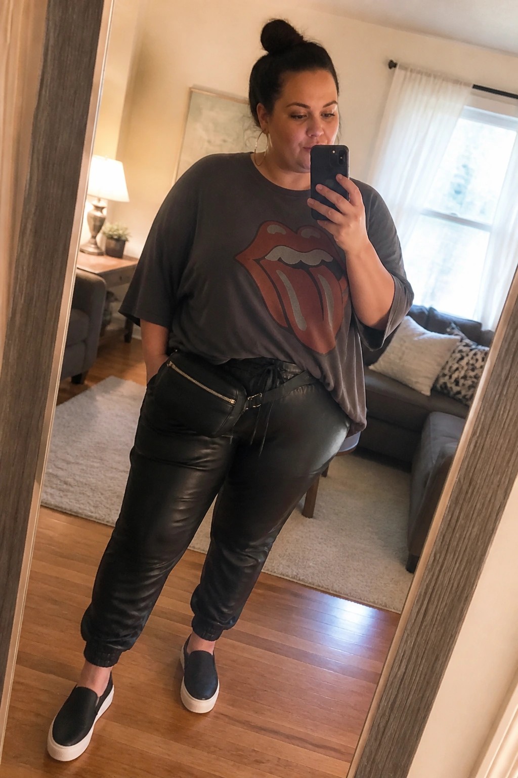 Plus size woman wearing an oversized gray Rolling Stones graphic tee tucked into black faux leather joggers with an attached belt bag and black slip on sneakers while taking a mirror selfie.