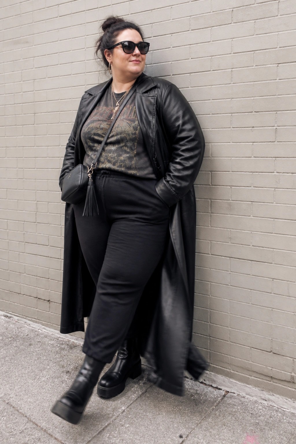 Woman in black leather coat, sunglasses, and platform boots leaning on brick wall