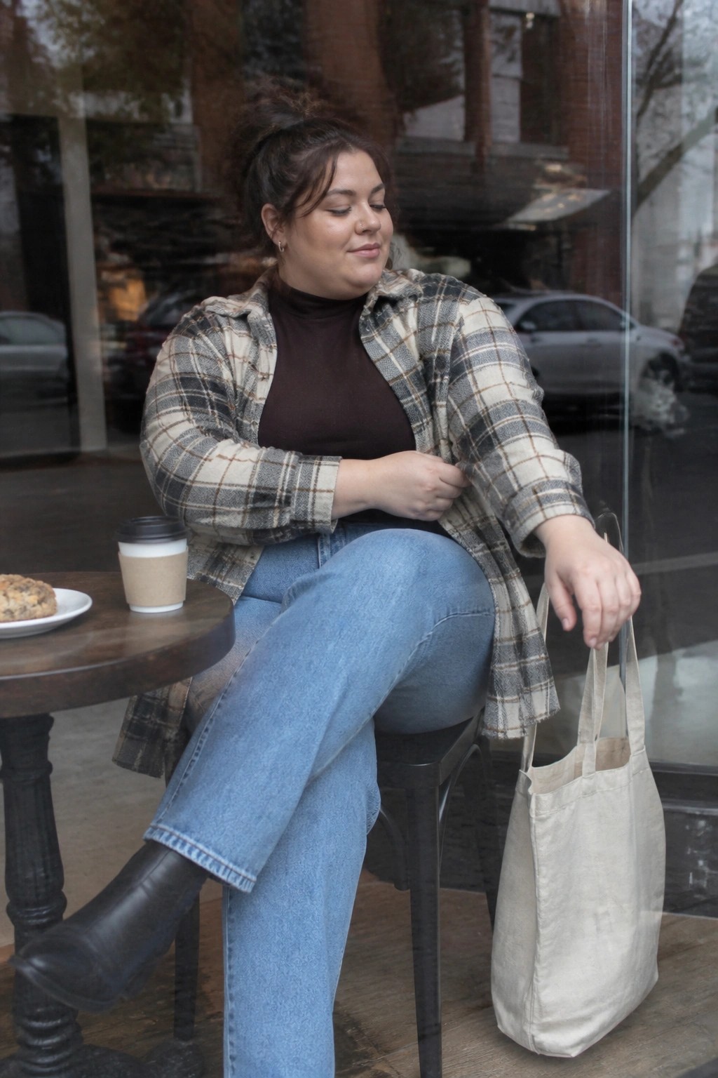 Woman in plaid shirt and jeans seated outdoors holding tote bag by cafe table