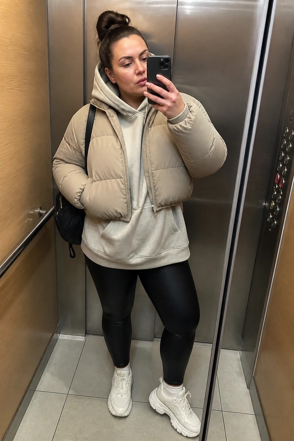 Woman in beige puffer vest and black leggings taking elevator selfie