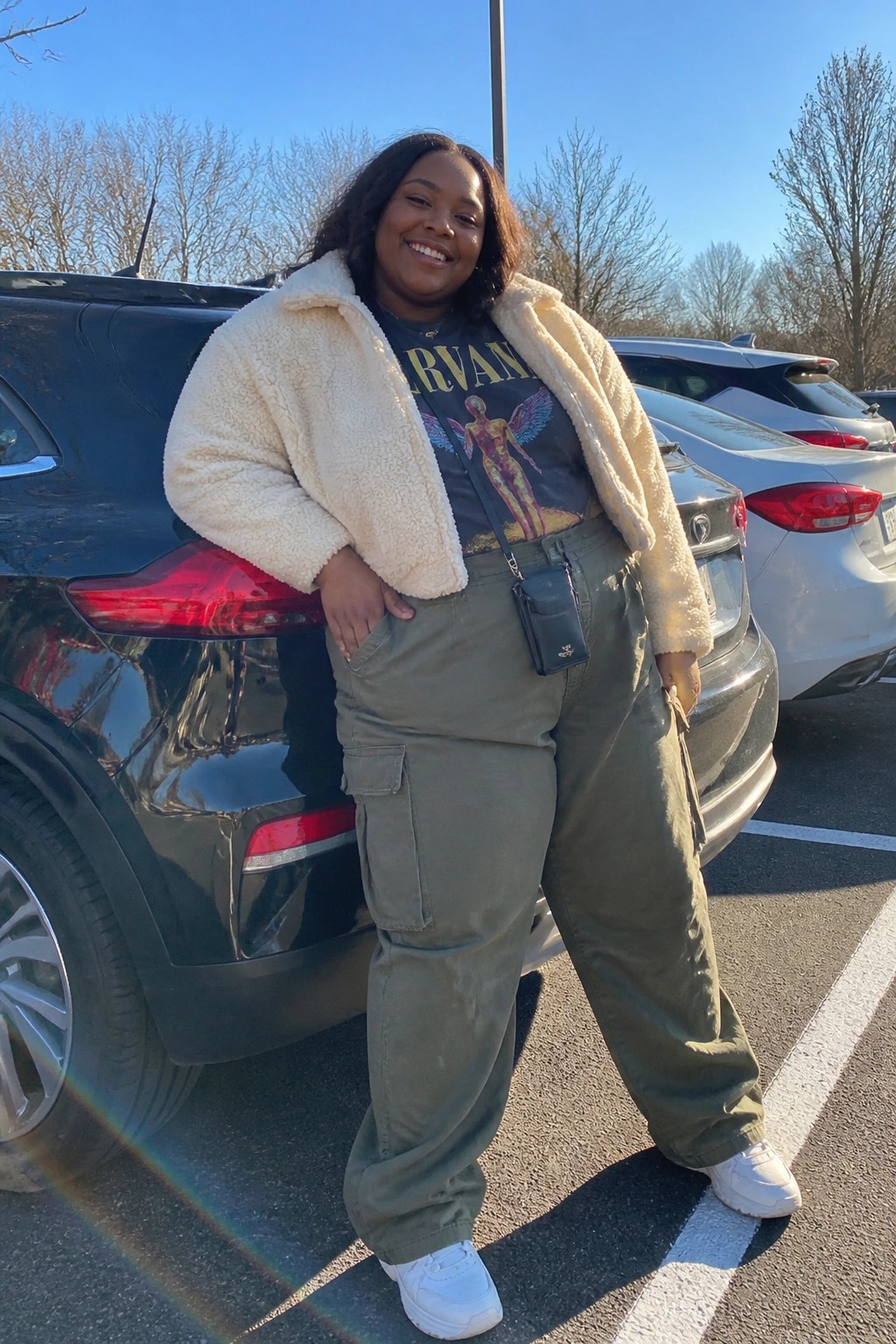 Woman in cream sherpa jacket and olive cargo pants smiling beside black SUV