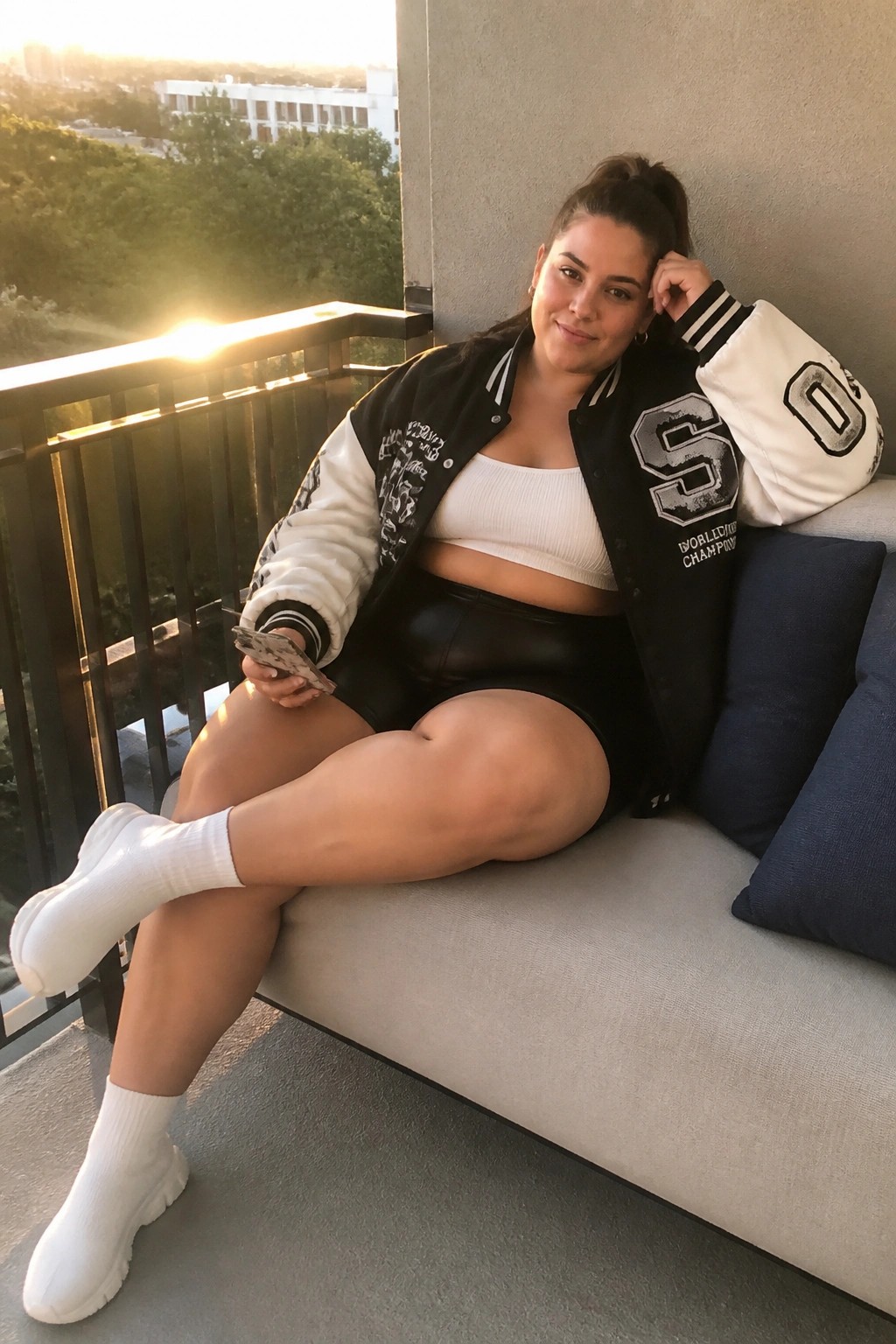 Woman in varsity jacket and leather shorts relaxes on balcony couch at sunset