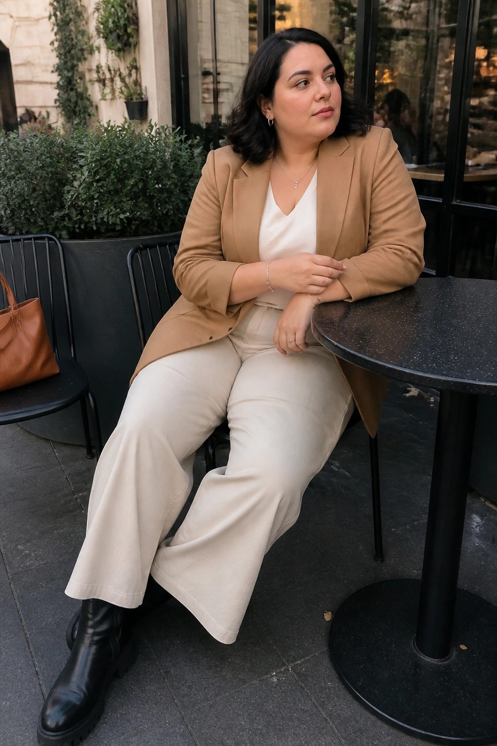 Woman in tan blazer and beige pants seated at outdoor table with brown bag nearby