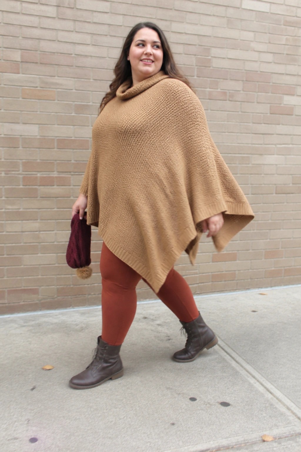 Woman in tan knit poncho and rust leggings holding maroon beanie by brick wall