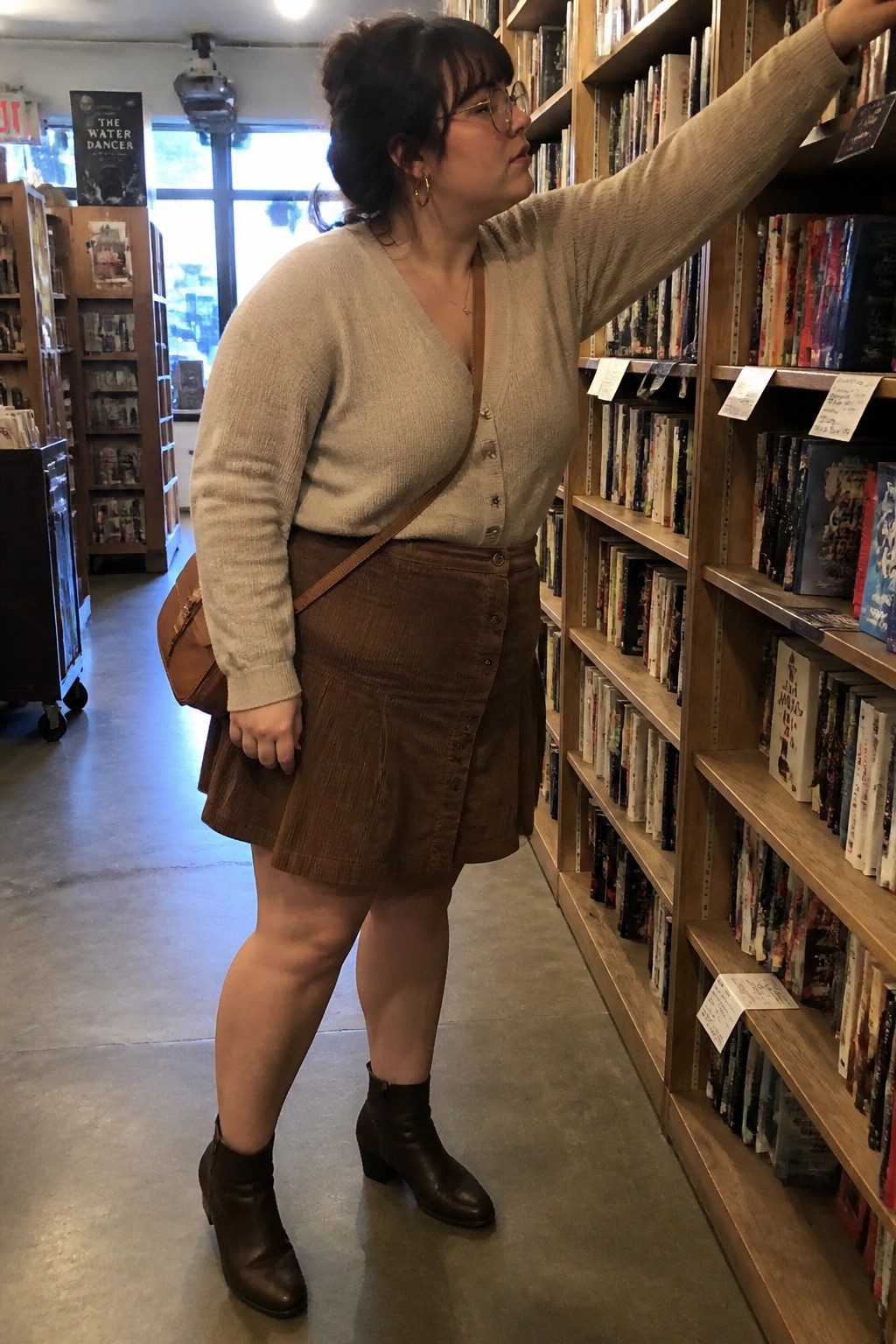 Woman in beige cardigan and brown corduroy skirt reaches for book on shelf.