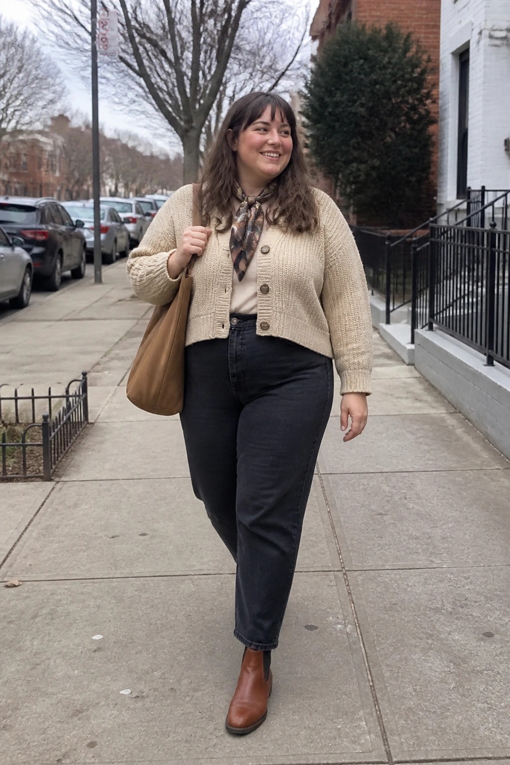 Smiling woman in cream cardigan and jeans carrying brown tote on sidewalk