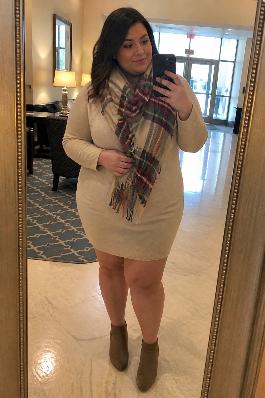 Woman in beige sweater dress with plaid scarf taking mirror selfie in lobby
