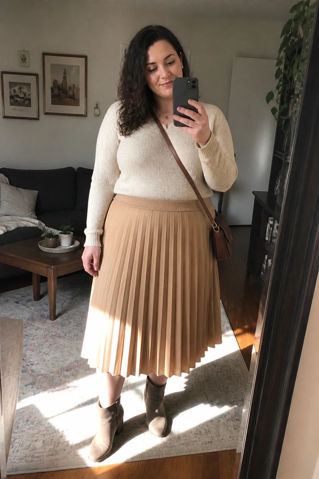 Woman in cream sweater and tan pleated skirt taking mirror selfie in living room