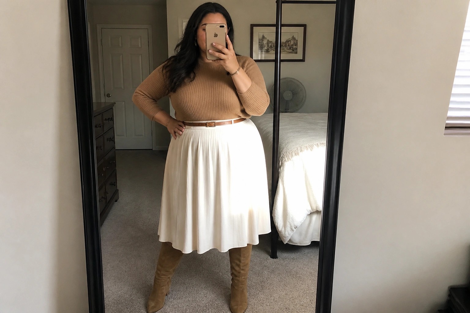 Plus size woman wearing a tan ribbed knit sweater tucked into a cream midi skirt with a brown belt and matching tan knee high boots taking a mirror selfie in a bedroom.