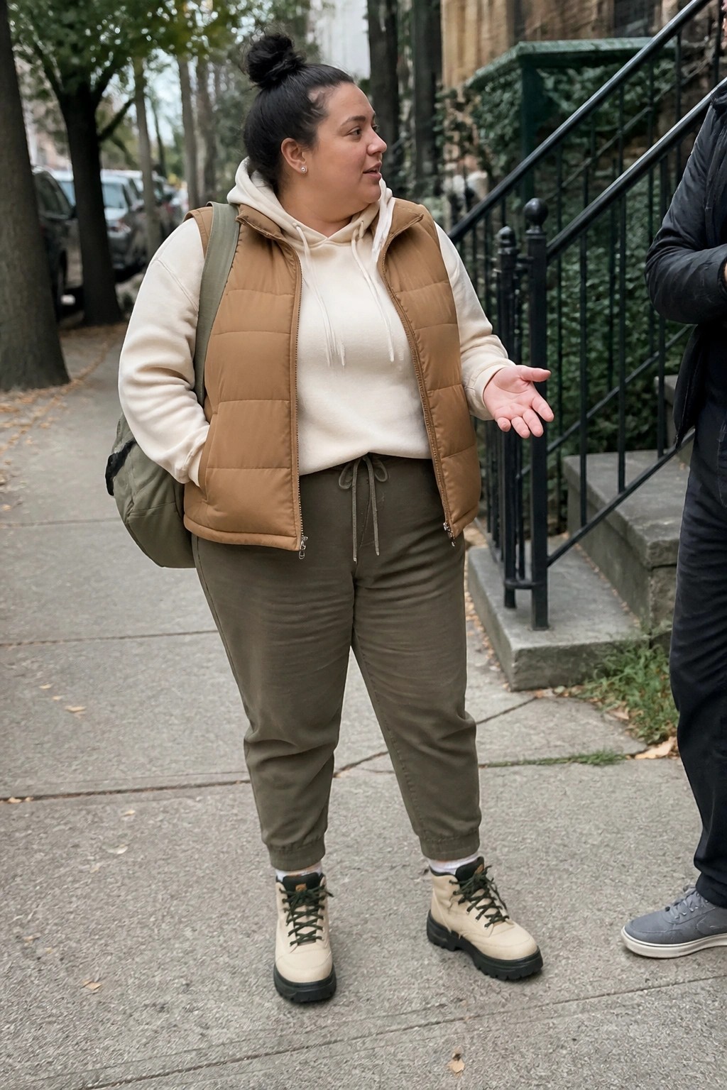Woman in cream hoodie and brown puffer vest gesturing on city sidewalk