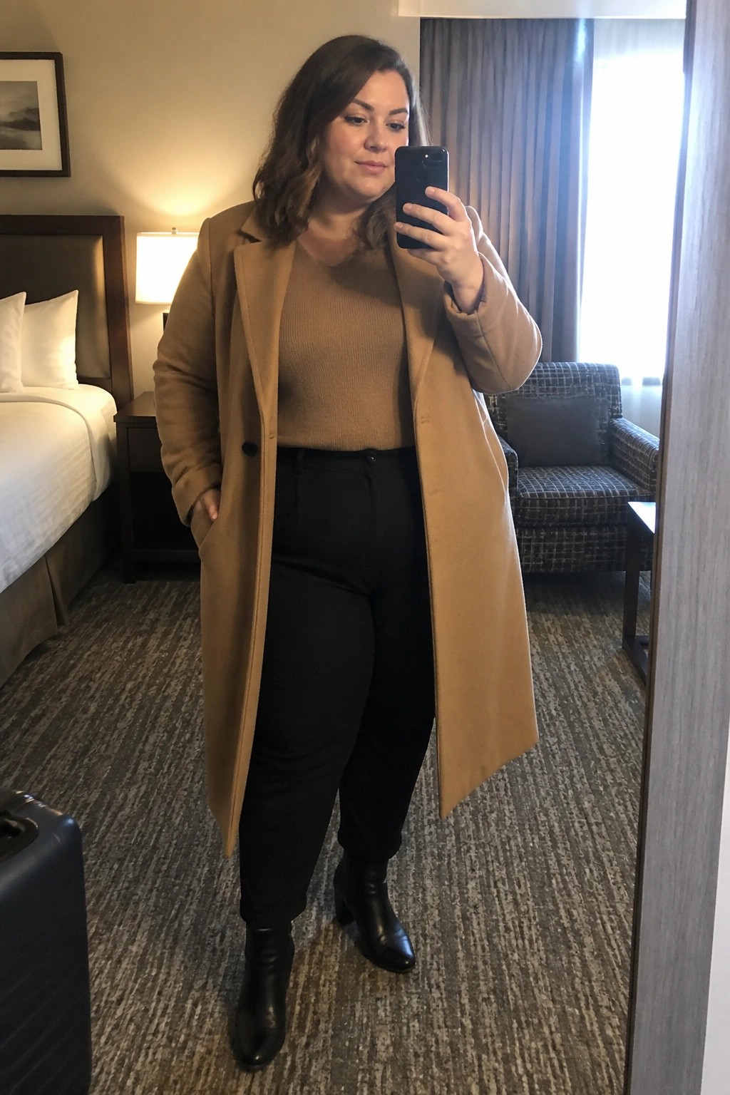 Woman in tan coat over brown sweater taking mirror selfie in hotel room