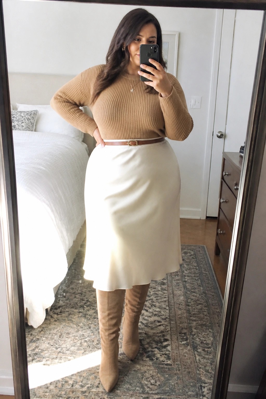 Plus size woman wearing a tan ribbed knit sweater tucked into a cream midi skirt with a brown belt and matching tan knee high boots taking a mirror selfie in a bedroom.