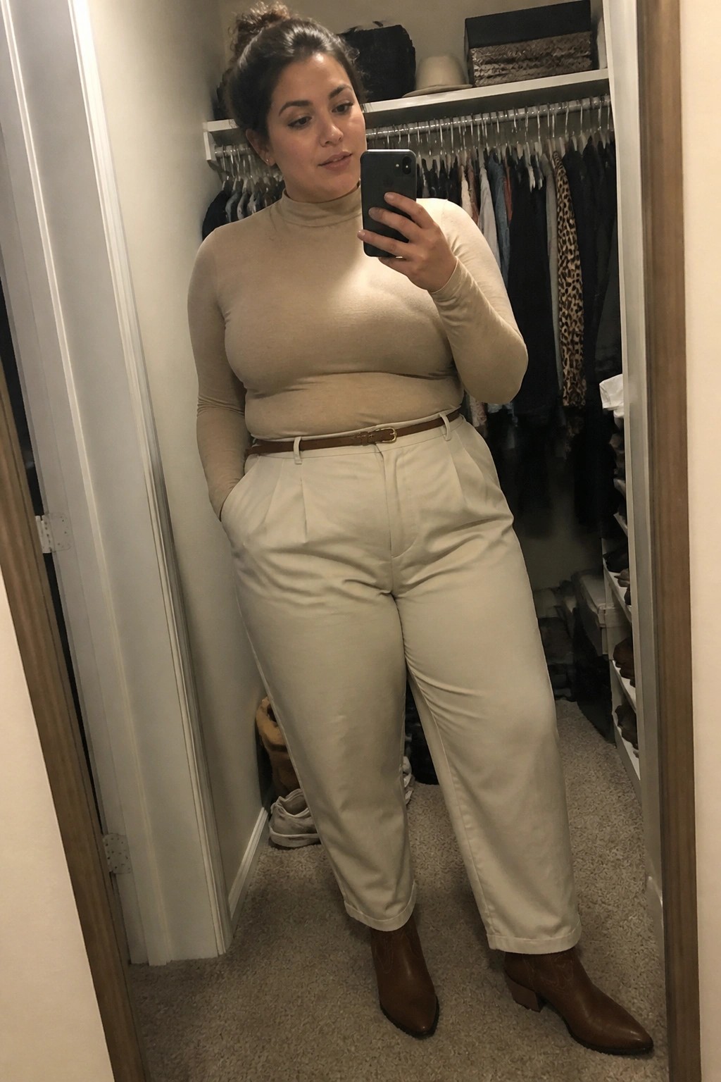 Woman in beige turtleneck and cream pants taking mirror selfie in closet.