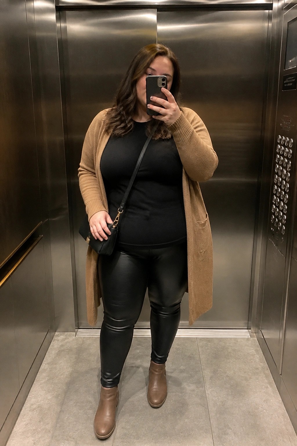 Plus size woman wearing a long tan open cardigan over a black top and black shiny leggings with matching tan ankle boots and a black crossbody bag while taking a mirror selfie in an elevator.