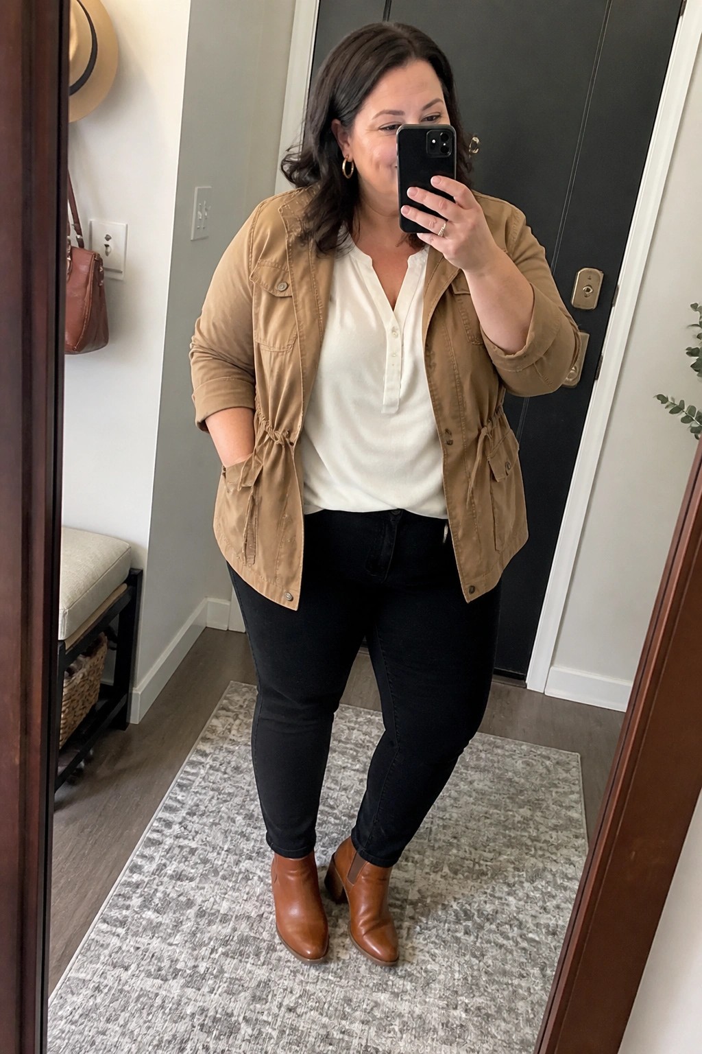 Woman in tan utility jacket and black jeans taking mirror selfie indoors