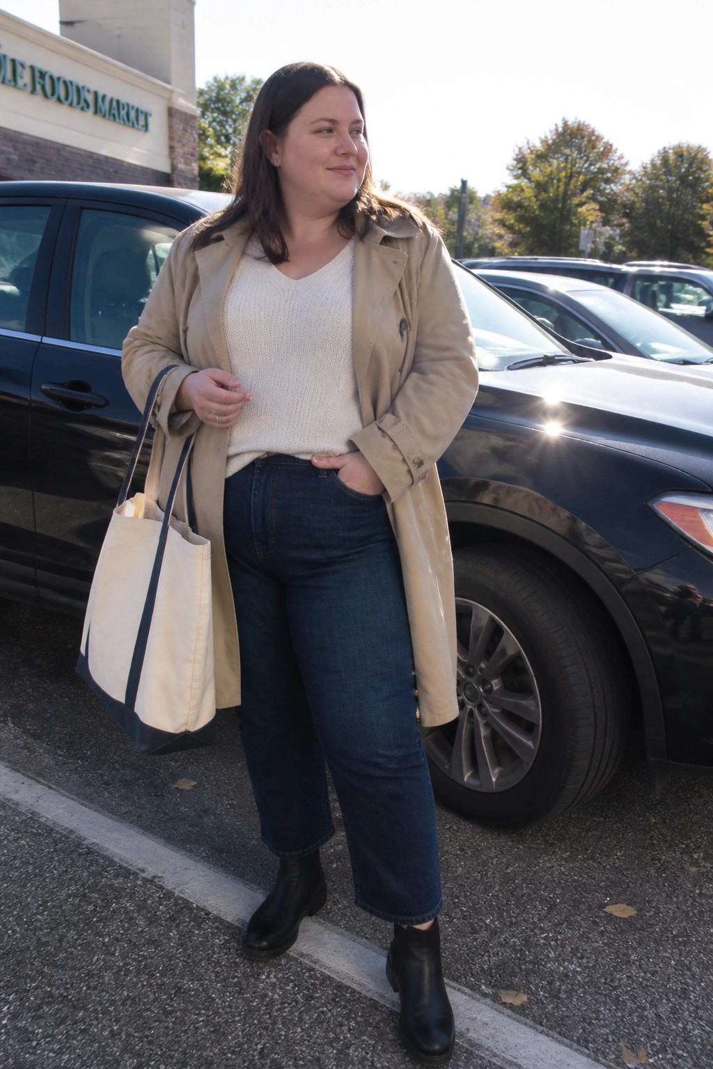 Plus size woman in beige trench coat layered over cream sweater with blue wide leg jeans and black ankle boots holding a canvas tote bag.