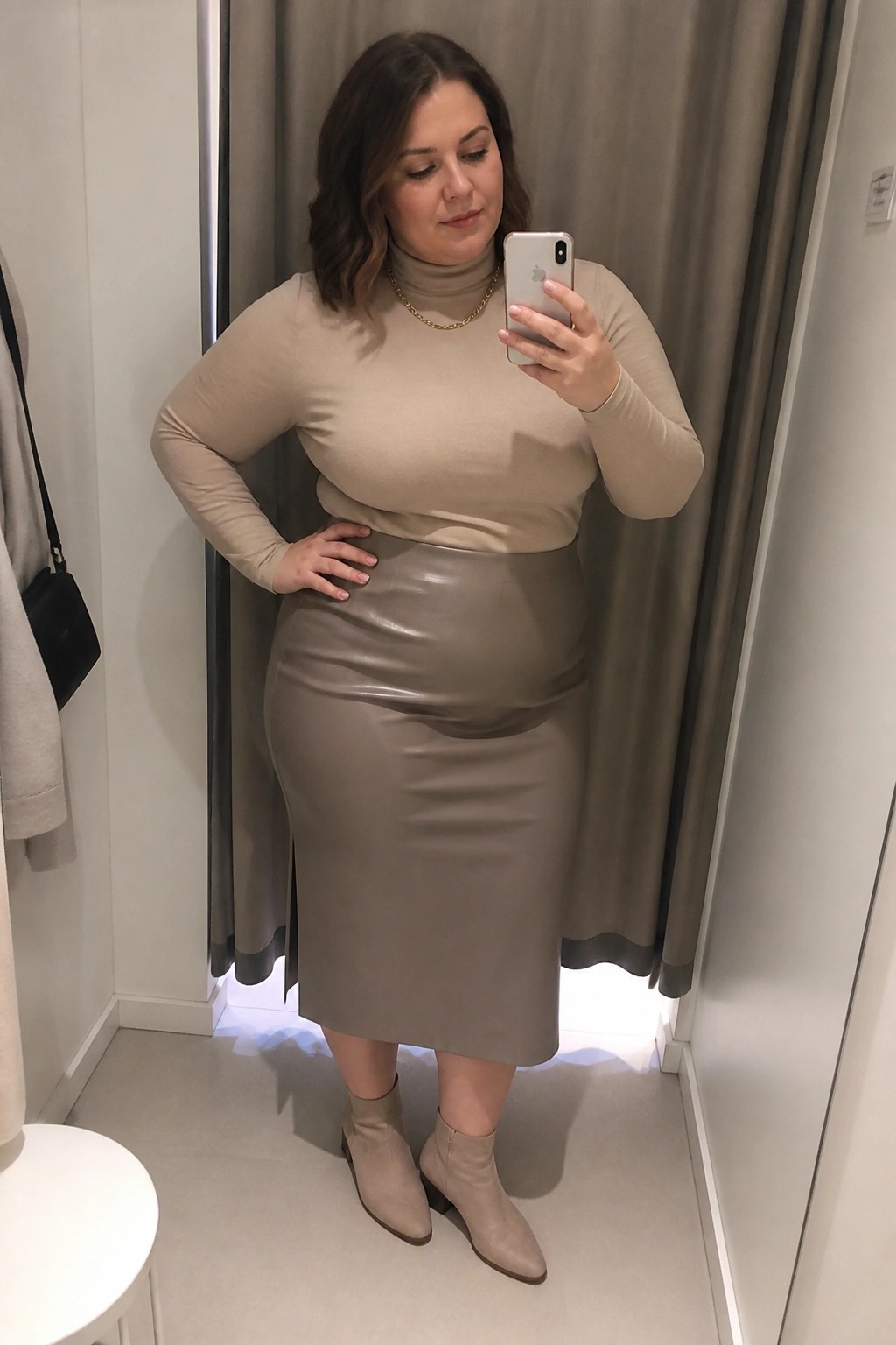 Plus size woman wearing a beige turtleneck top tucked into a taupe leather midi skirt with matching ankle boots and a gold necklace while taking a mirror selfie in a fitting room.