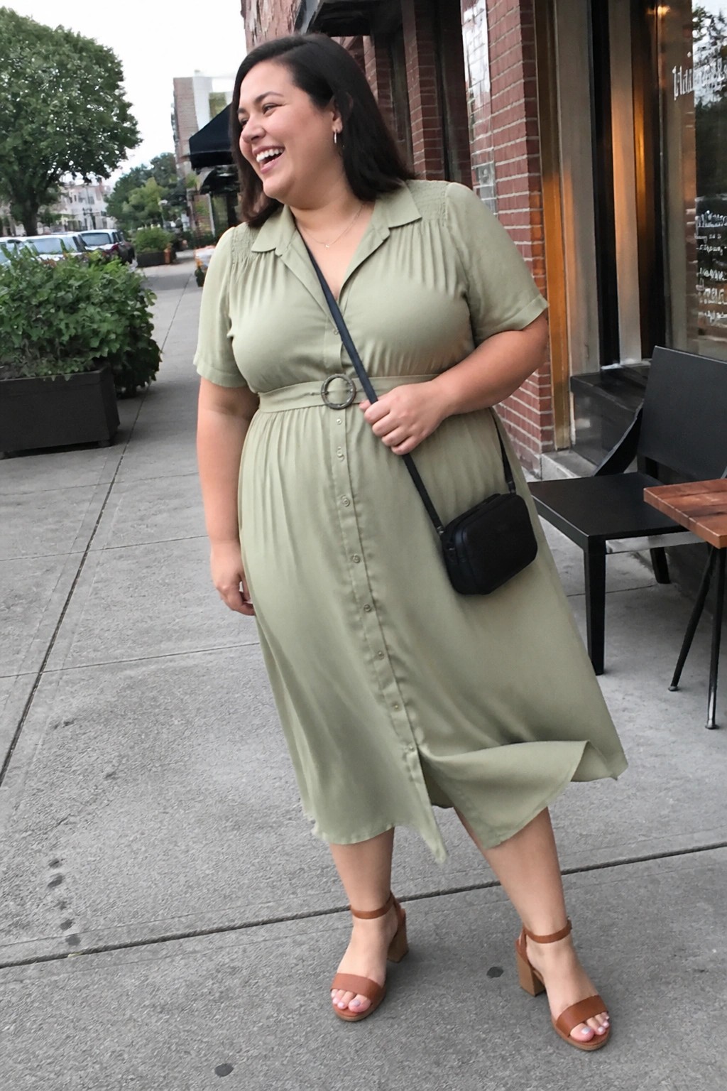 Smiling woman in sage green midi dress with black bag on city sidewalk.