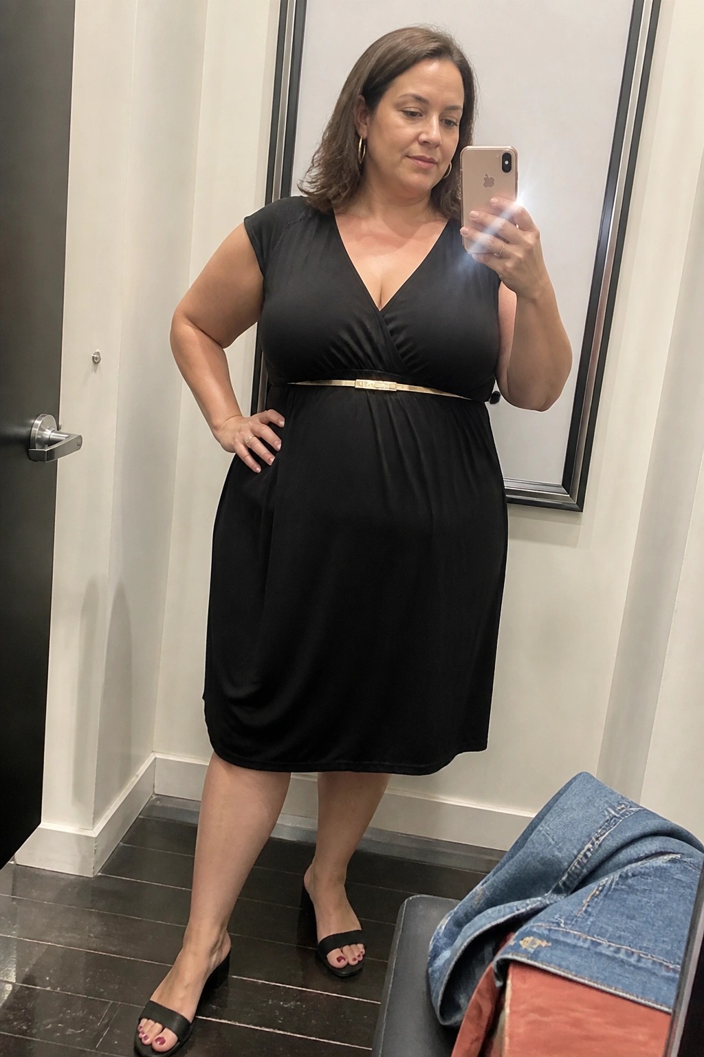 Woman wearing a plus size black sleeveless wrap dress with thin gold belt and black heeled sandals taking a mirror selfie in a fitting room.