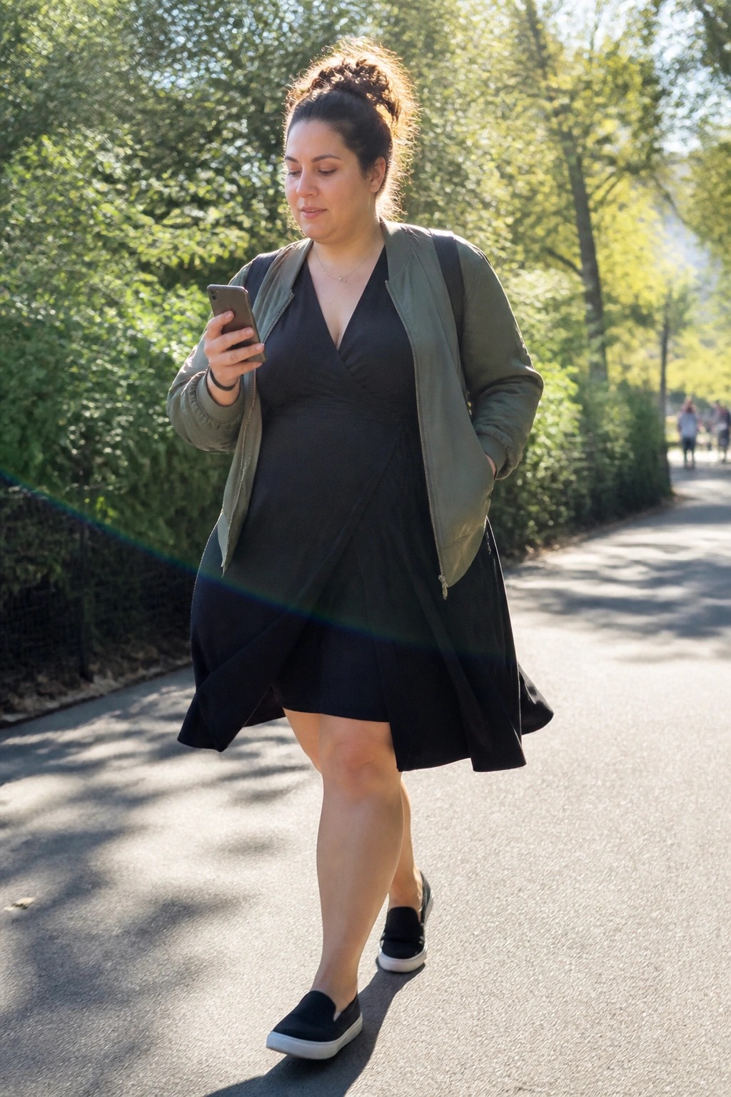 Woman in black dress and green jacket checks phone on sunny park path.