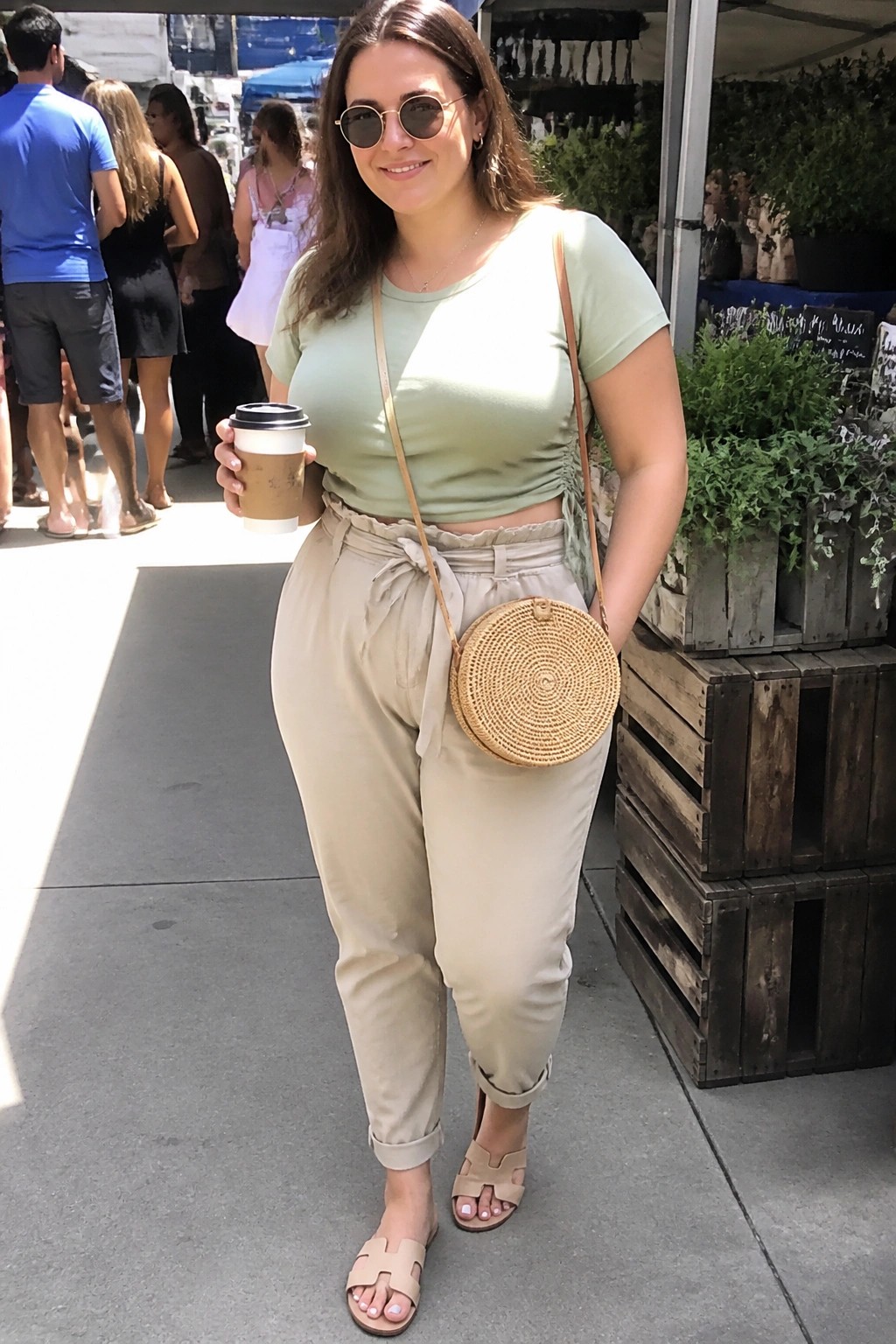 Woman in sage green crop top and beige pants holding coffee at market