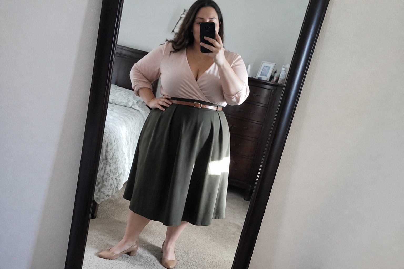 Woman in spotted top and green skirt taking bedroom mirror selfie with phone