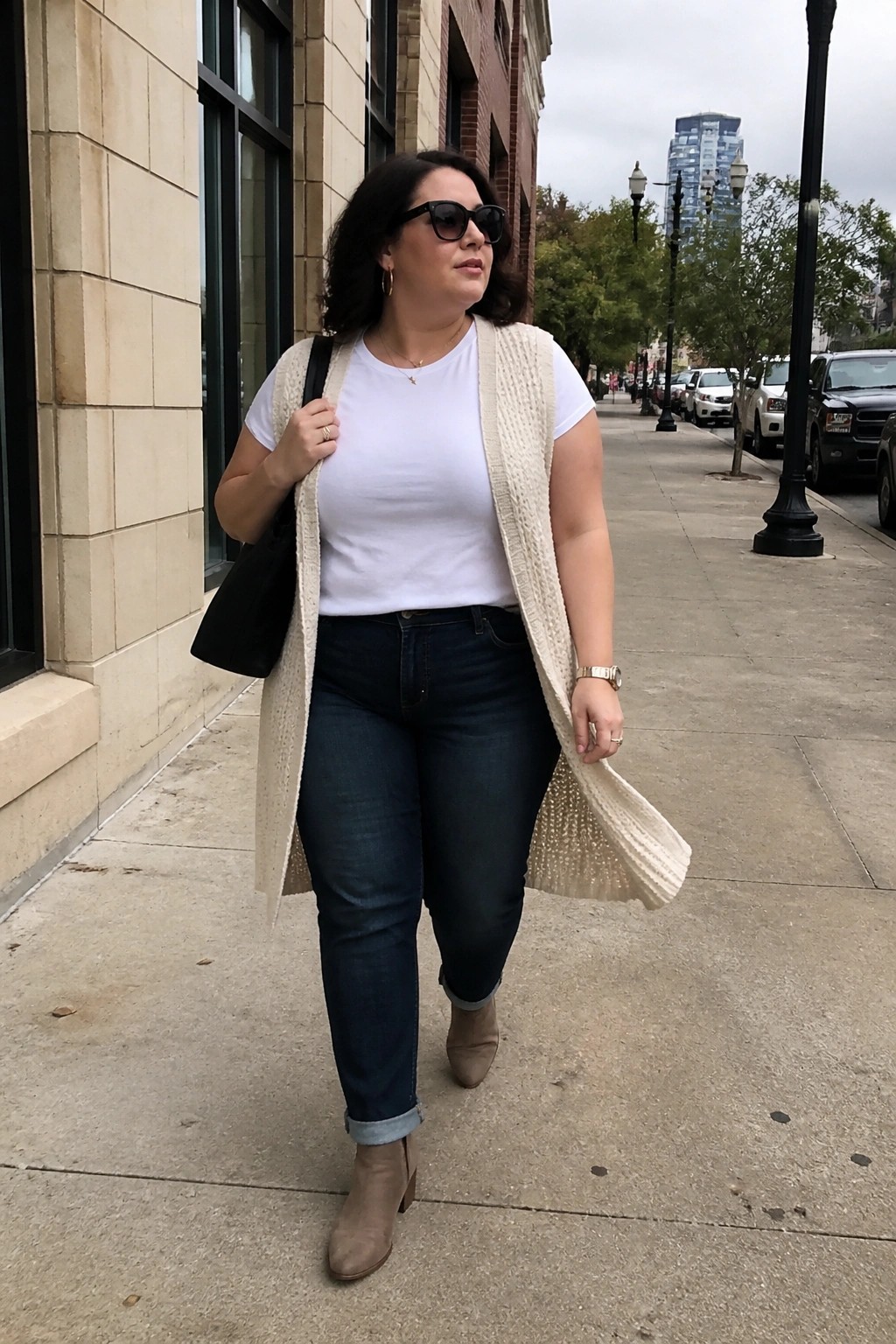 Woman in cream knit vest, white tee, and jeans walking on city sidewalk