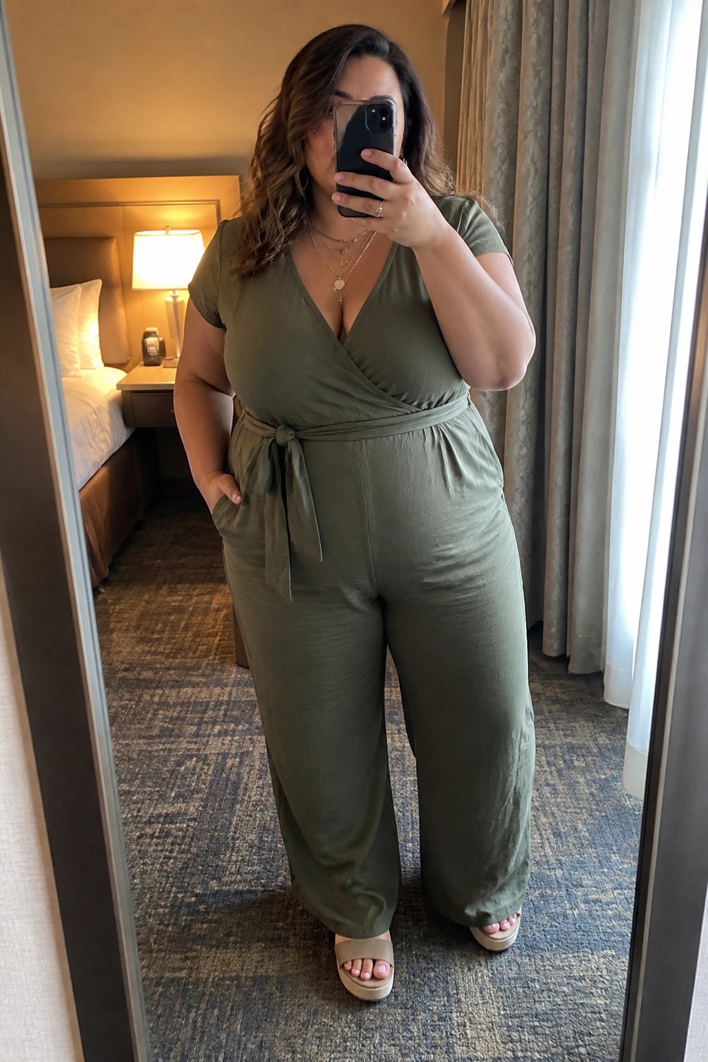 Woman in olive green jumpsuit taking mirror selfie in hotel room
