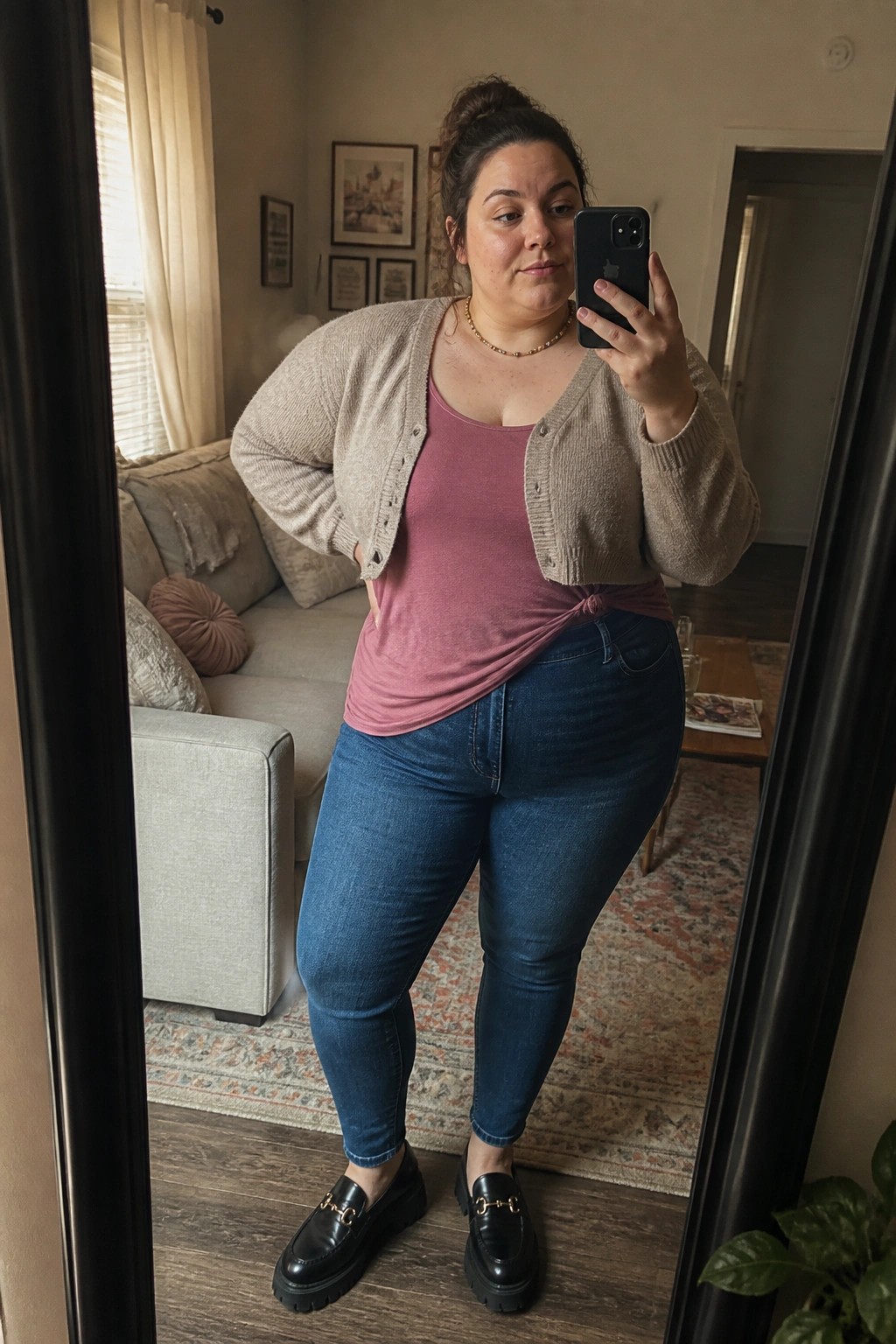 Plus size woman in beige open cardigan over pink knotted tank top blue jeans and black loafers posing for mirror selfie.