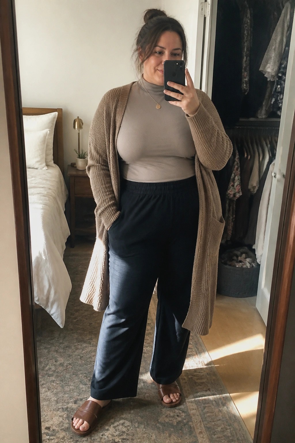 Woman in beige cardigan, gray top, and black pants taking bedroom mirror selfie