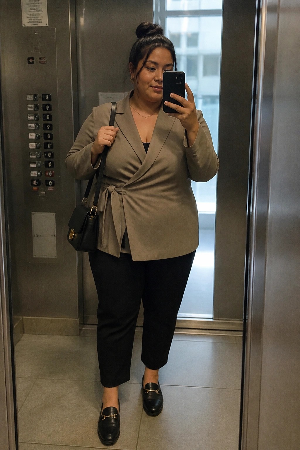 Woman in taupe wrap blazer taking mirror selfie in elevator with black bag