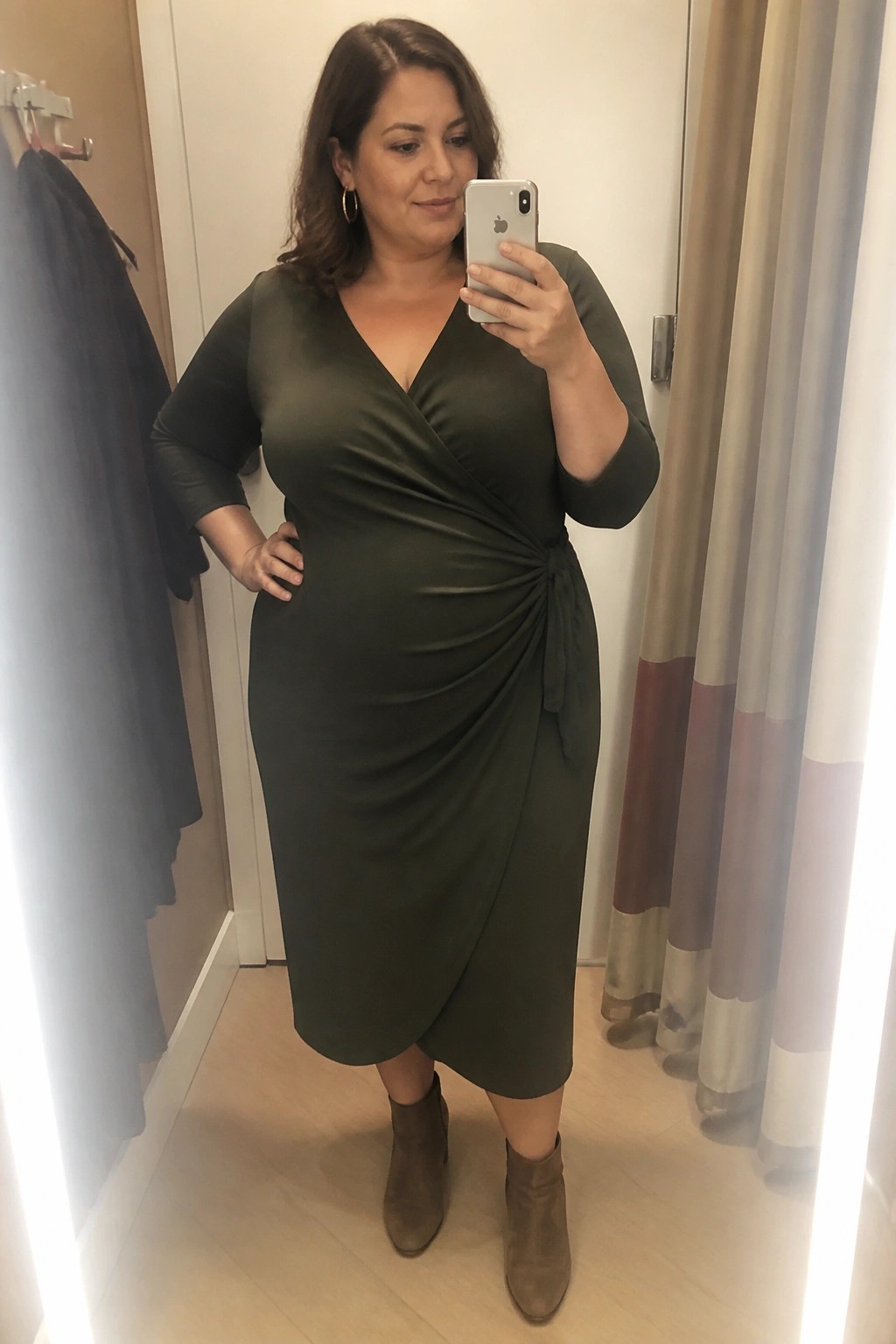 Woman in olive wrap dress and ankle boots taking mirror selfie with phone