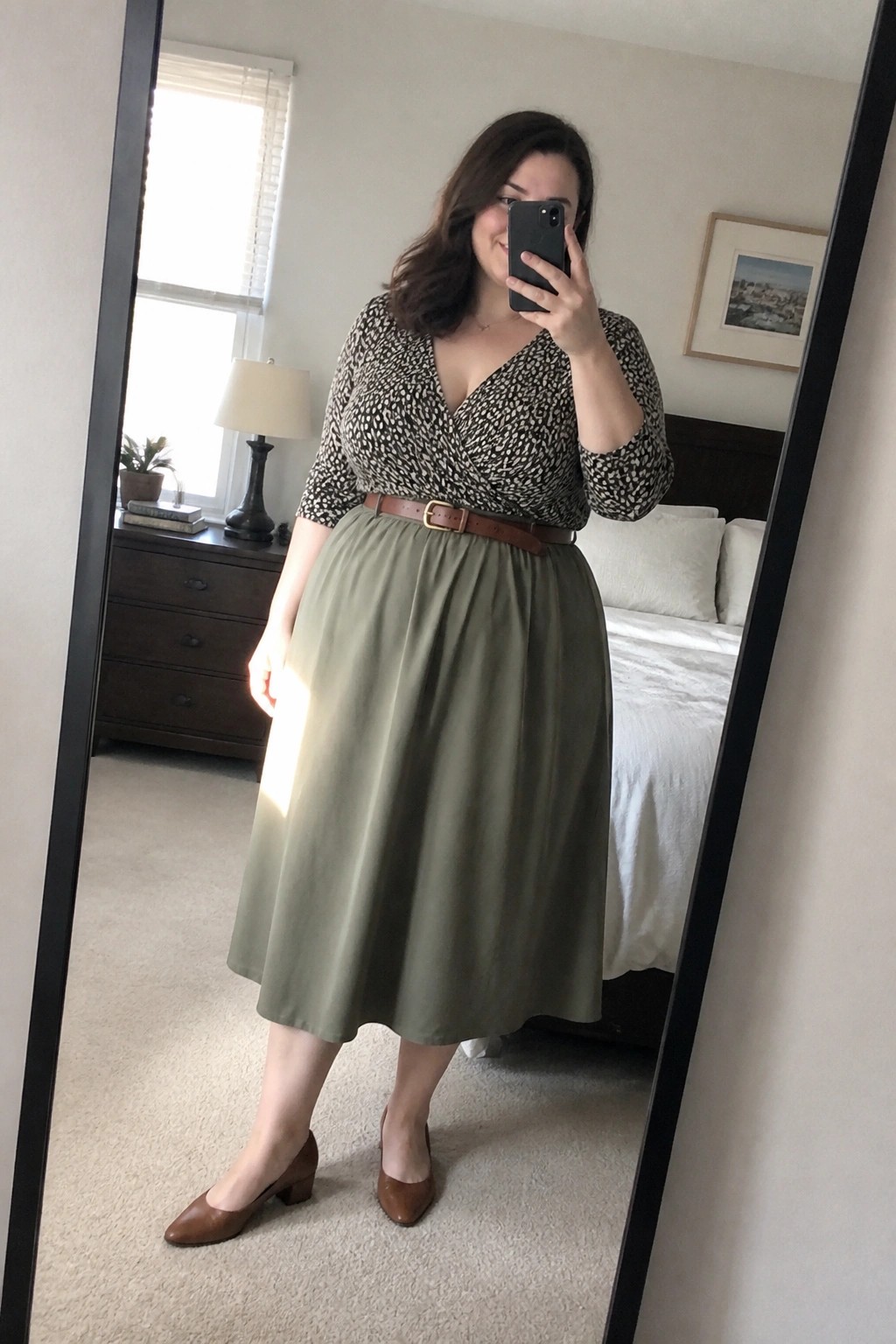Woman in spotted top and green skirt taking bedroom mirror selfie with phone