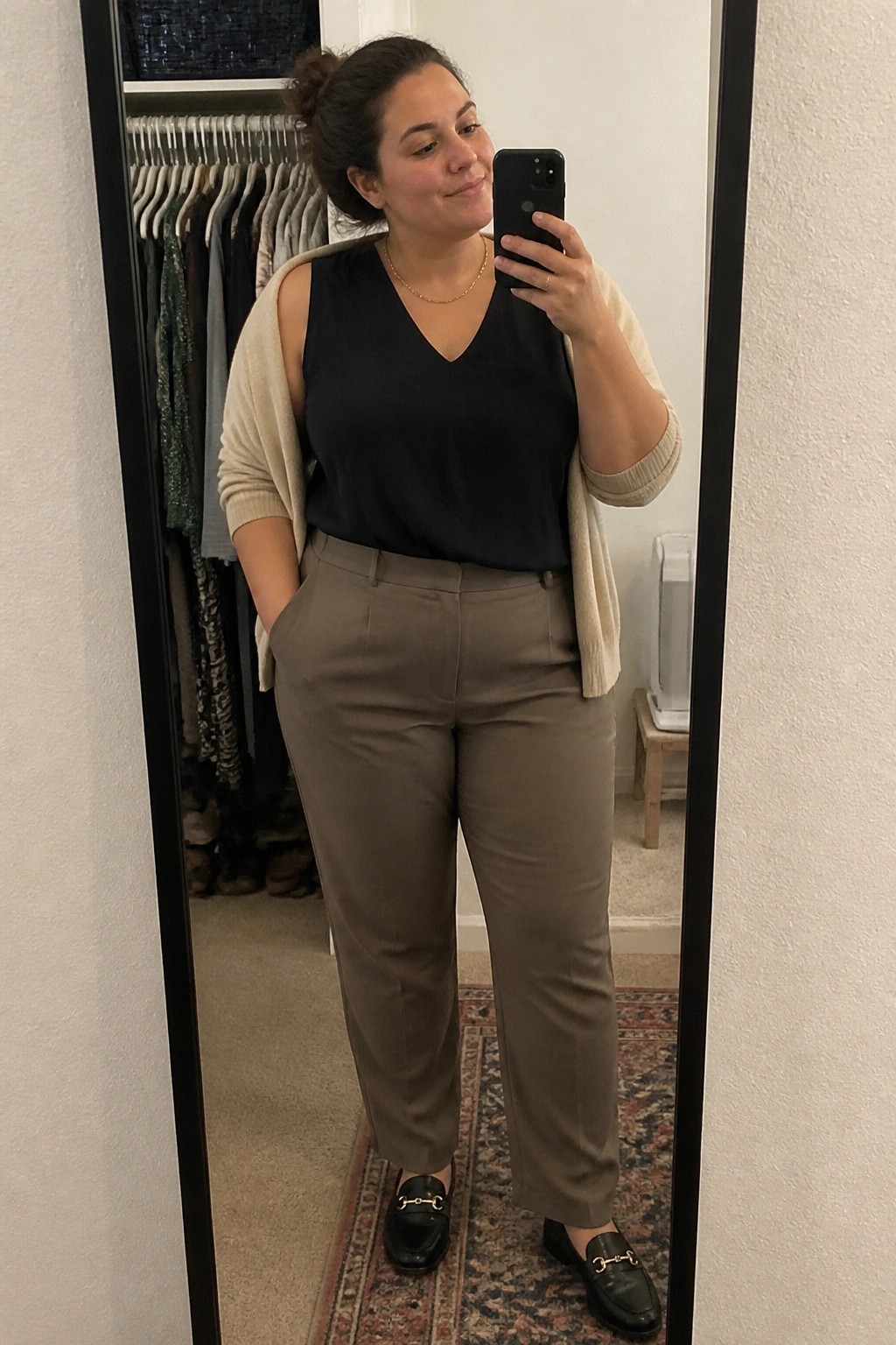 Woman in black top and beige cardigan taking mirror selfie in taupe pants