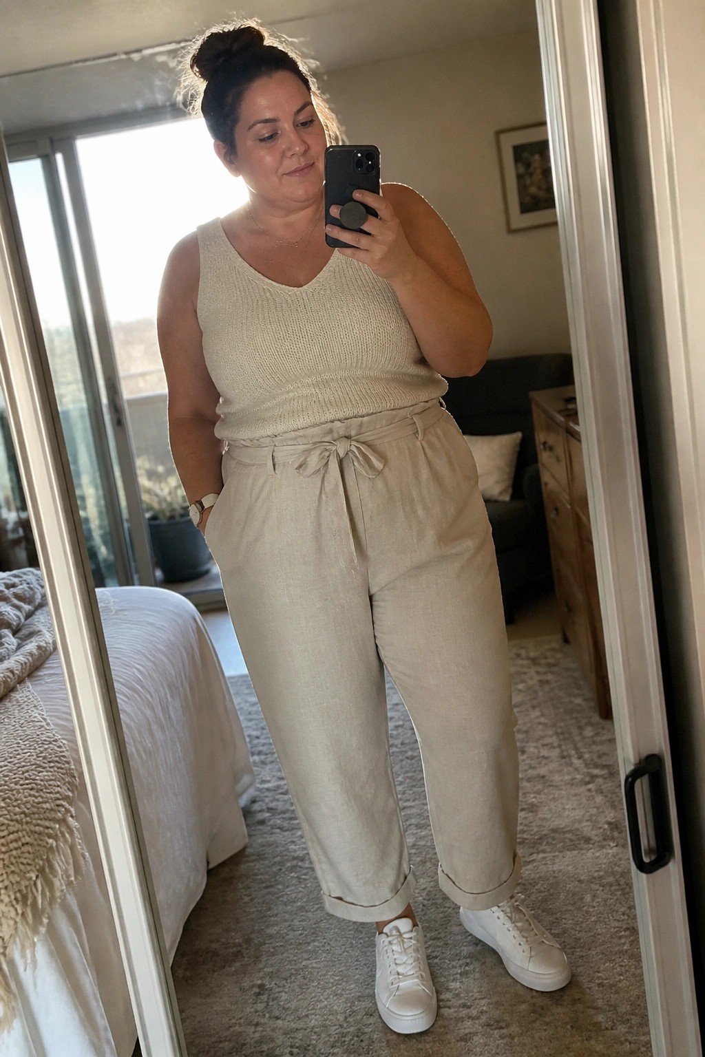 Woman in cream knit top and linen pants taking bedroom mirror selfie with phone