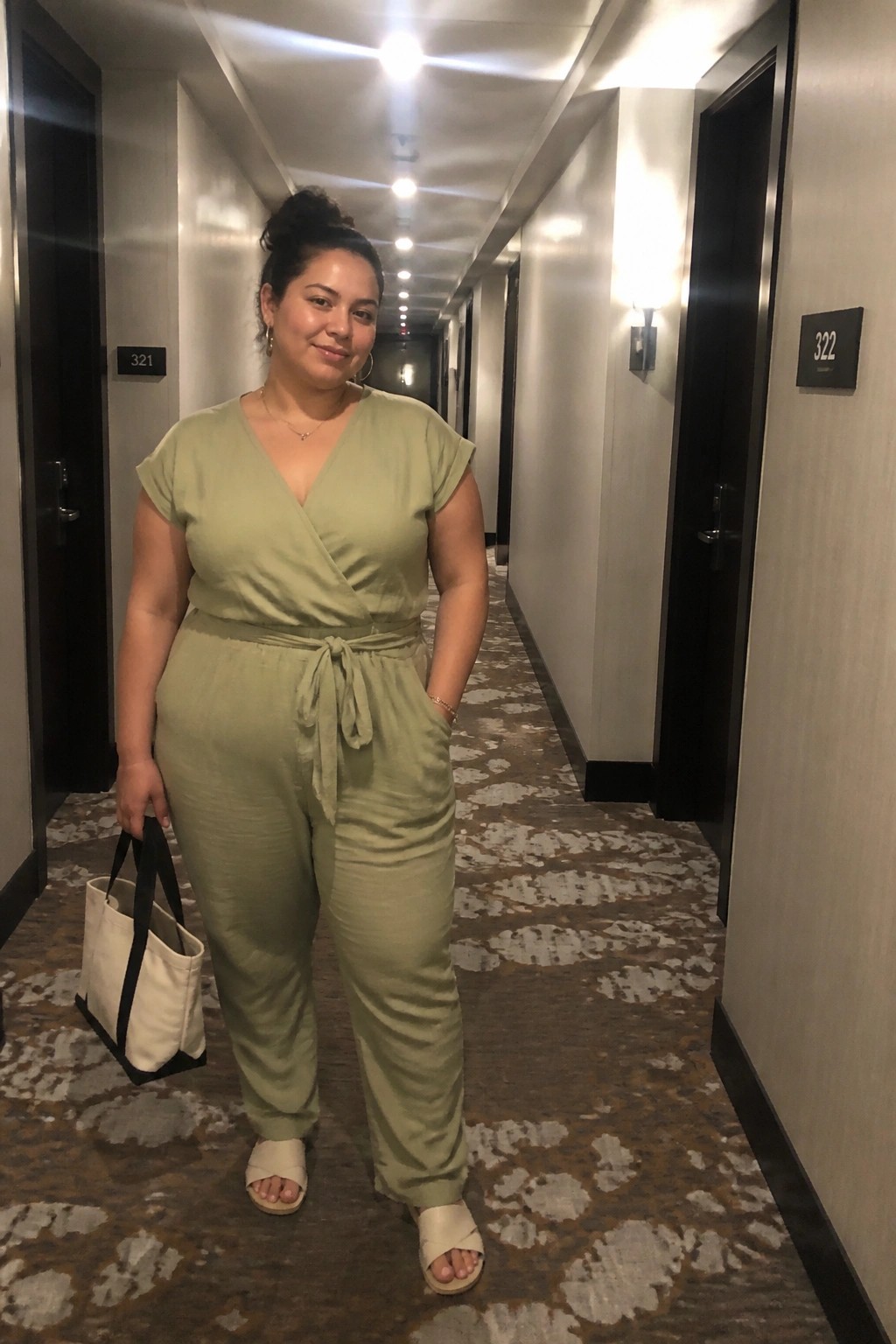 Woman in sage green jumpsuit holding white tote bag in hotel hallway.
