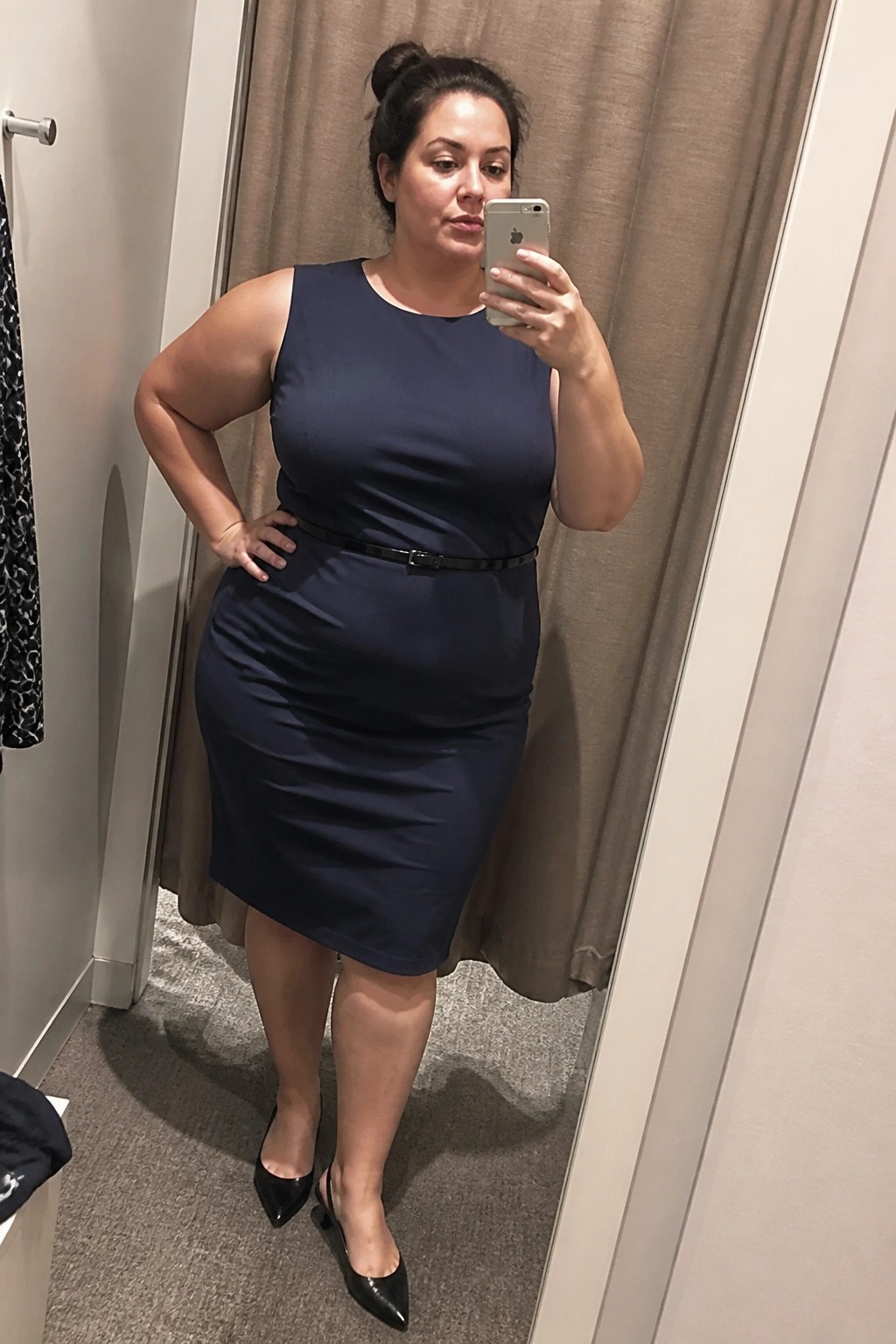 Plus size woman in navy sleeveless knee length sheath dress cinched with thin black belt and wearing black pointed heels while taking a mirror selfie in a fitting room.
