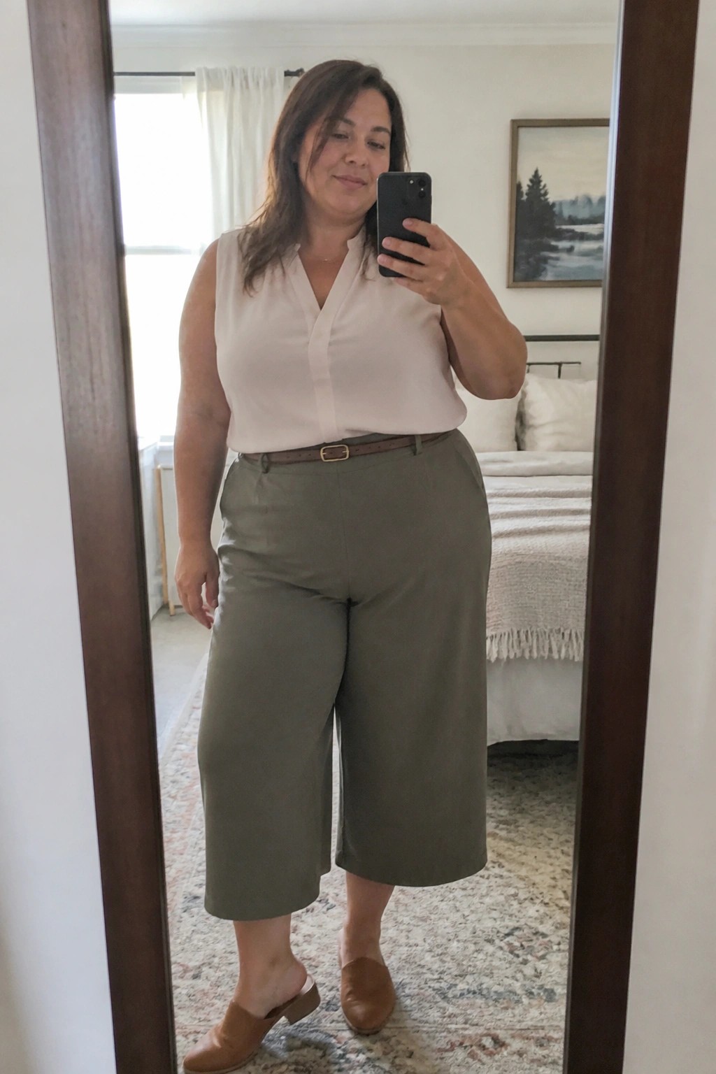 Woman in beige blouse and olive capri pants taking bedroom mirror selfie