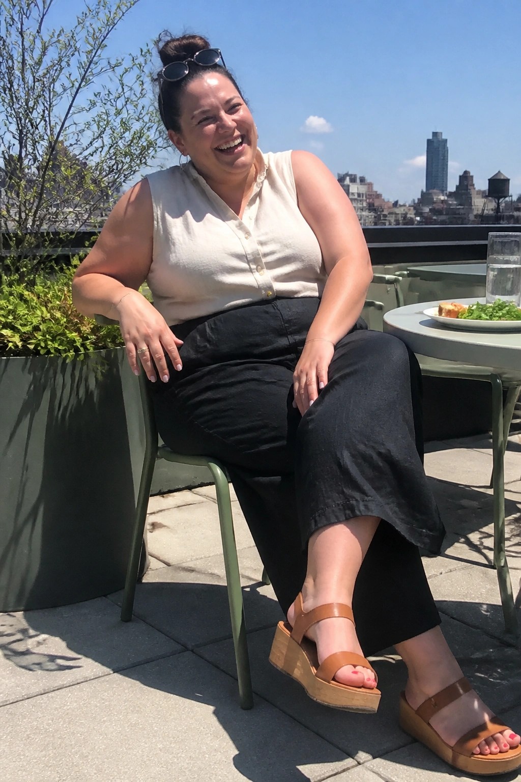 Smiling woman in beige sleeveless top and black pants on sunny city rooftop patio