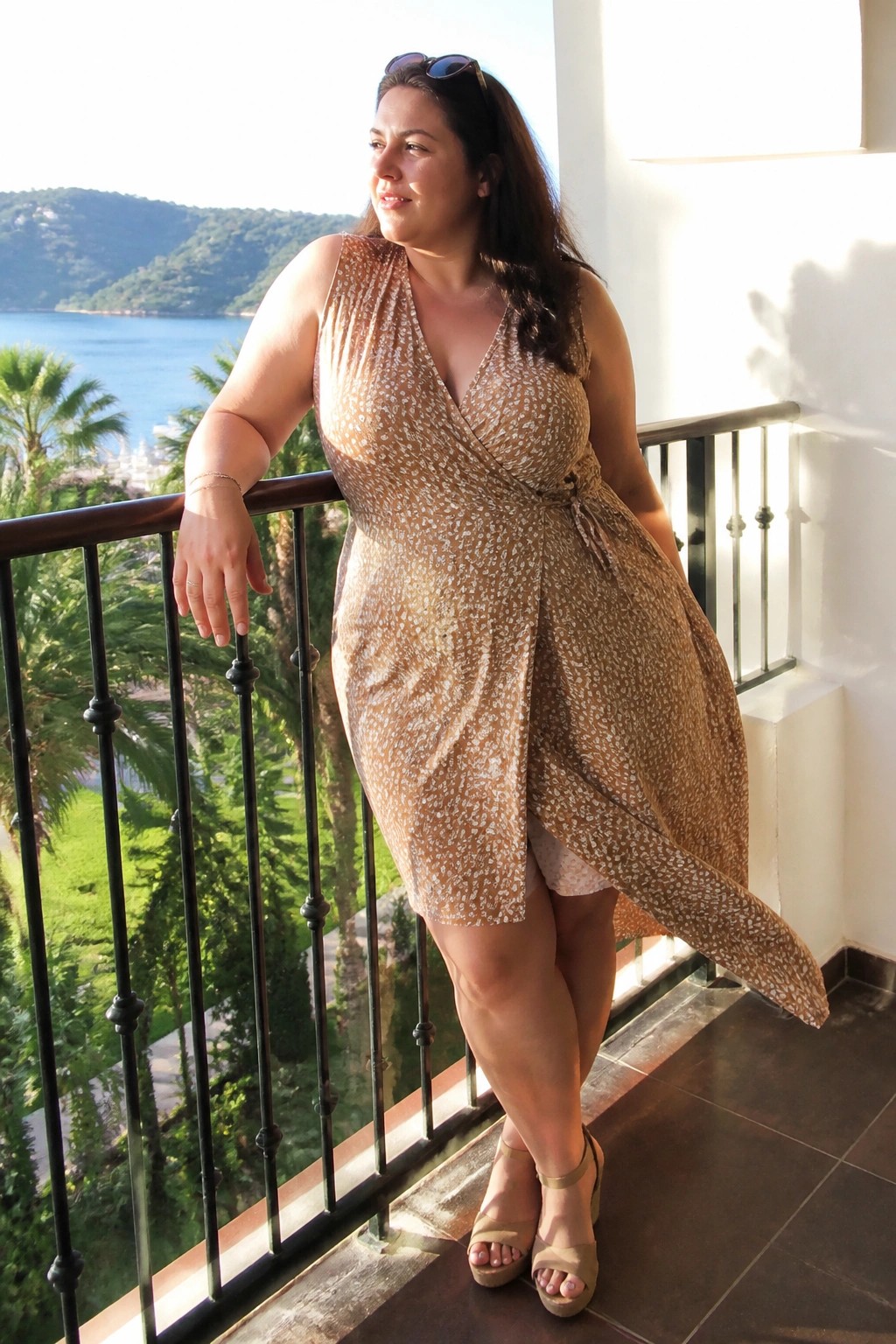 Plus size woman wearing a sleeveless tan wrap dress with white speckled print, tied at the waist, paired with beige wedge sandals, standing on a balcony.