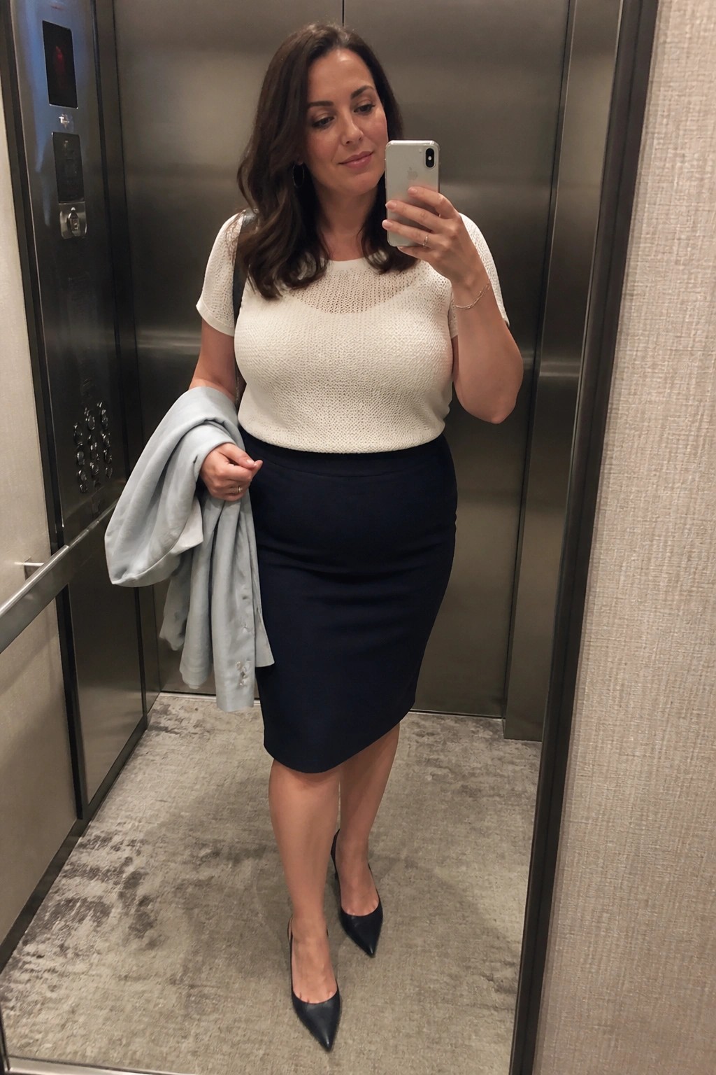 Woman in white knit top and navy skirt taking selfie in elevator holding blue jacket