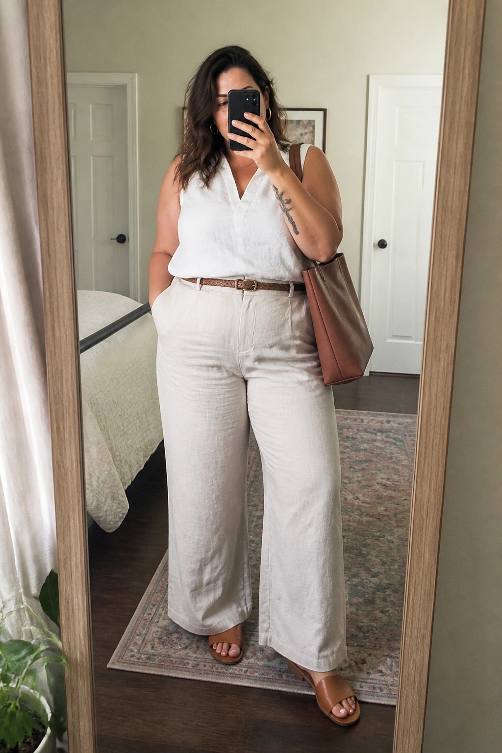 Woman in white sleeveless top and wide-leg pants taking mirror selfie with brown bag
