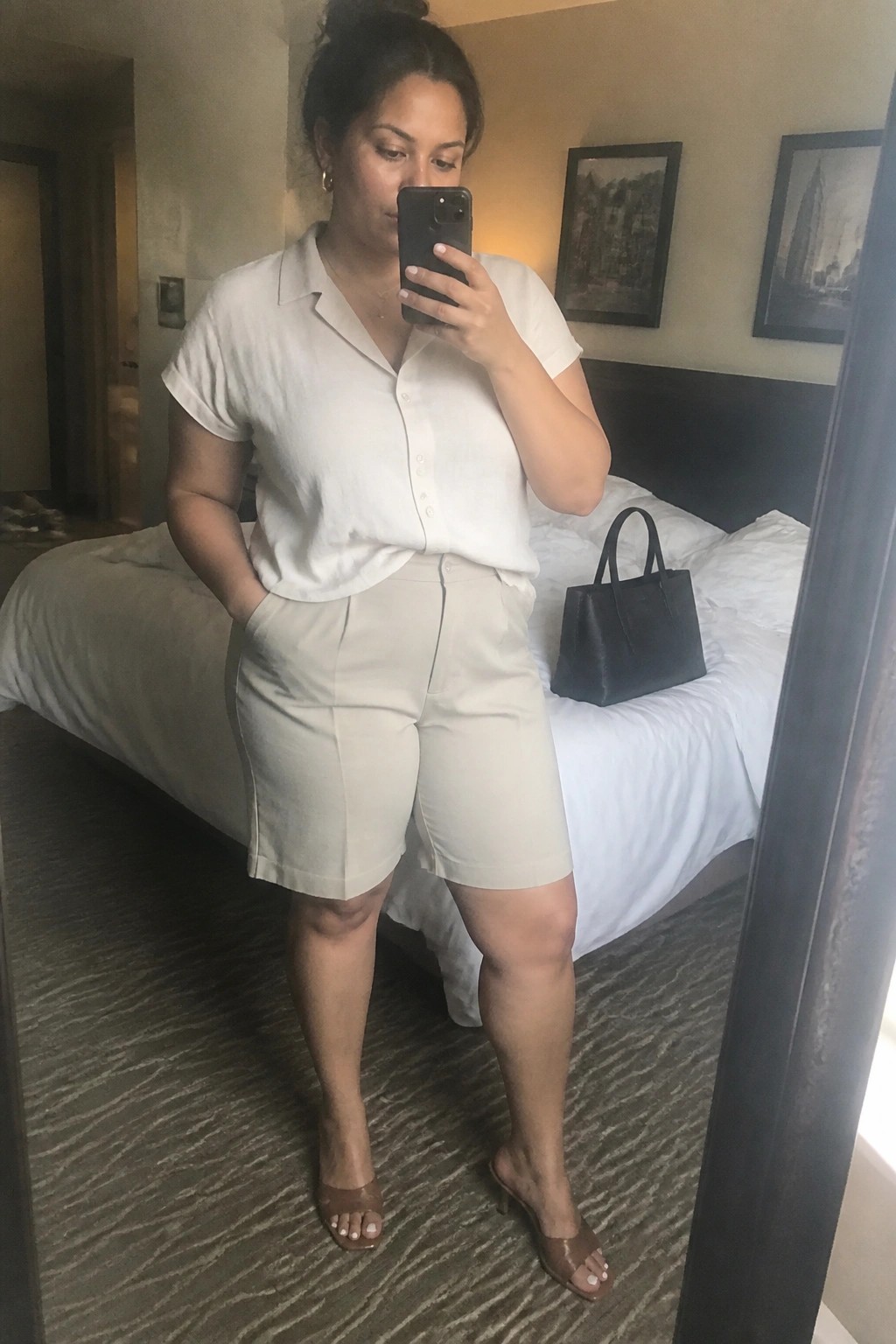 Woman in a white short sleeve button up shirt and beige Bermuda shorts taking a mirror selfie in a hotel room, wearing brown heeled mules with a black handbag visible on the bed behind her.