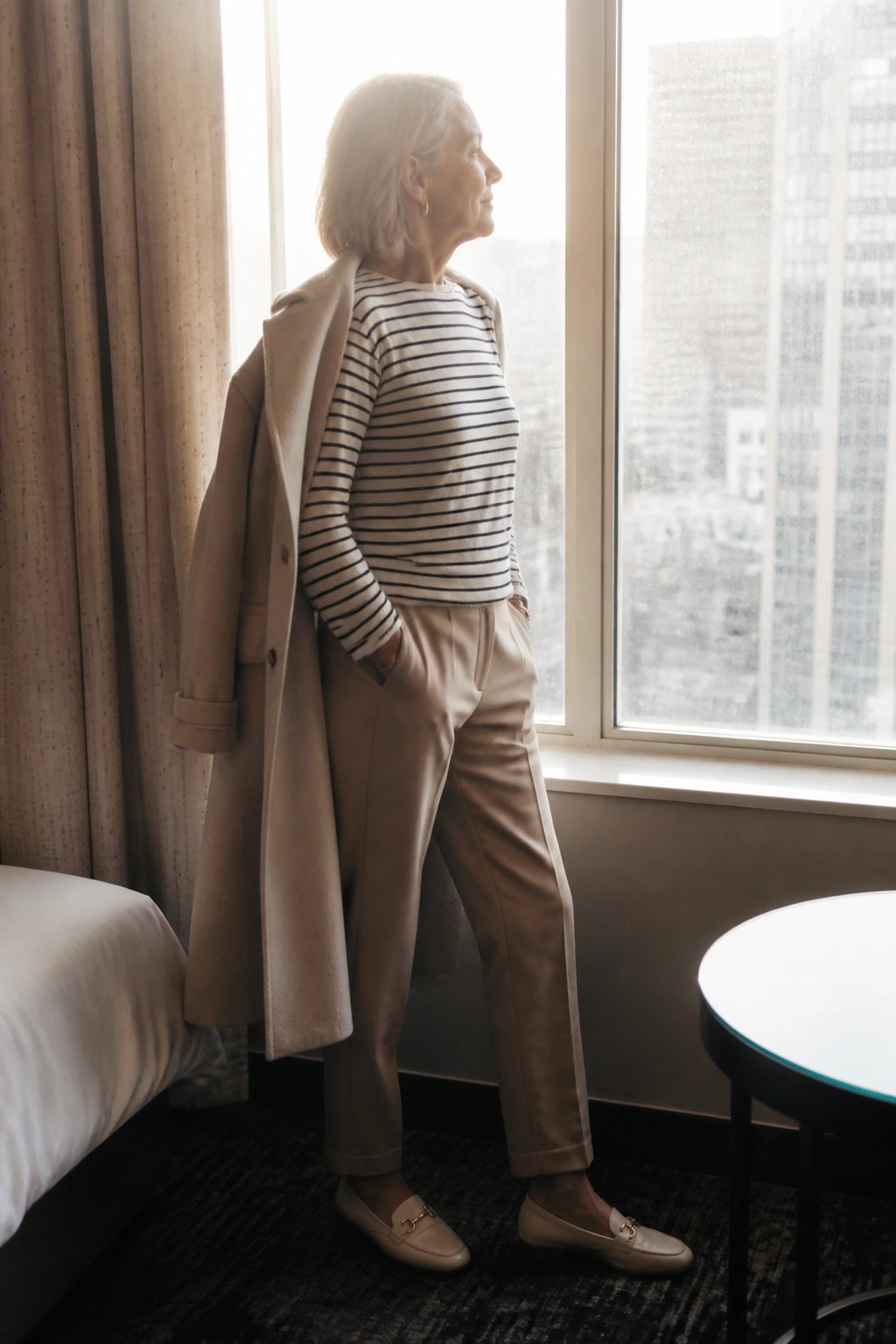 Older woman in striped shirt and beige coat gazes from hotel window.