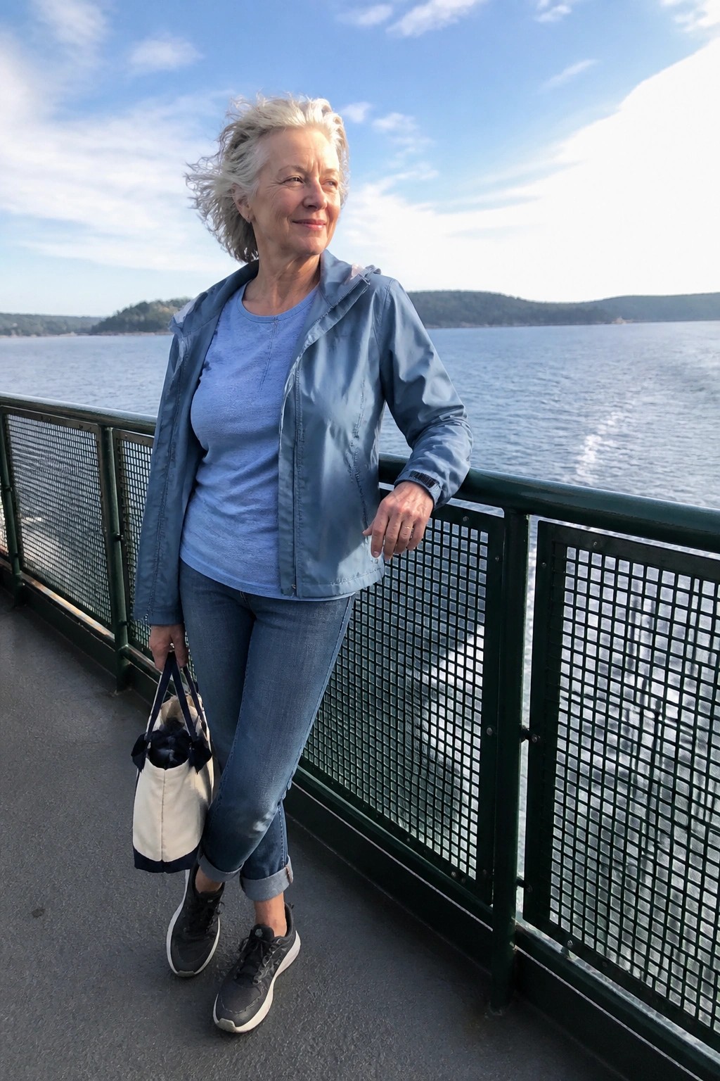Woman over 60 wearing a light blue zip jacket over a matching blue t-shirt with blue jeans cuffed at the ankles black sneakers and a cream tote bag standing on a deck by water.