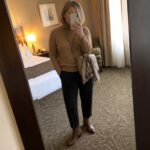 Woman in tan turtleneck sweater, black pants, brown boots taking mirror selfie in hotel room