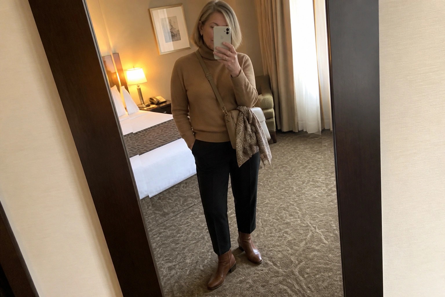 Woman in tan turtleneck sweater, black pants, brown boots taking mirror selfie in hotel room