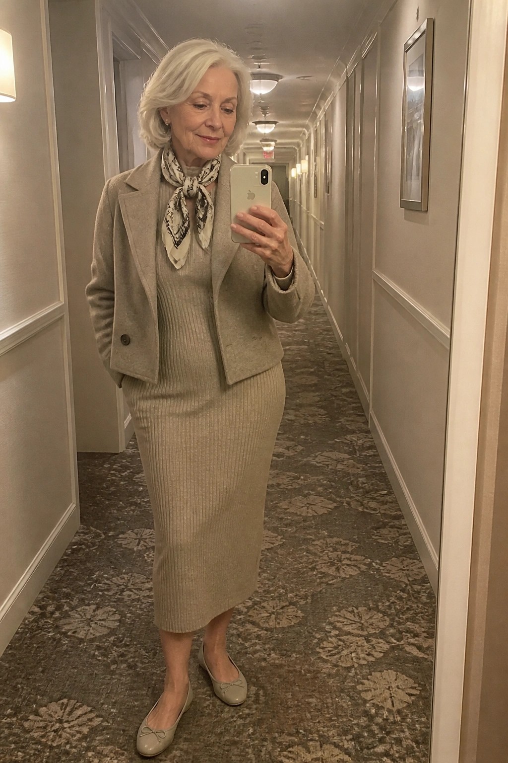 Woman over 60 wearing a taupe ribbed knit midi dress with matching cropped jacket, patterned scarf tied at the neck, and beige ballet flats while taking a mirror selfie in a hotel hallway.